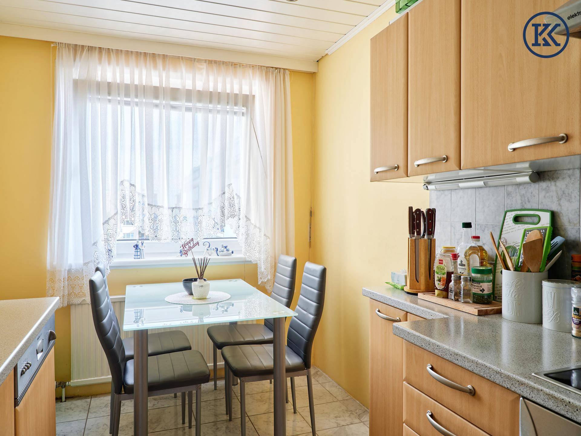 Helle Küche mit Essbereich, Holzschränken und Fenster mit Vorhängen.