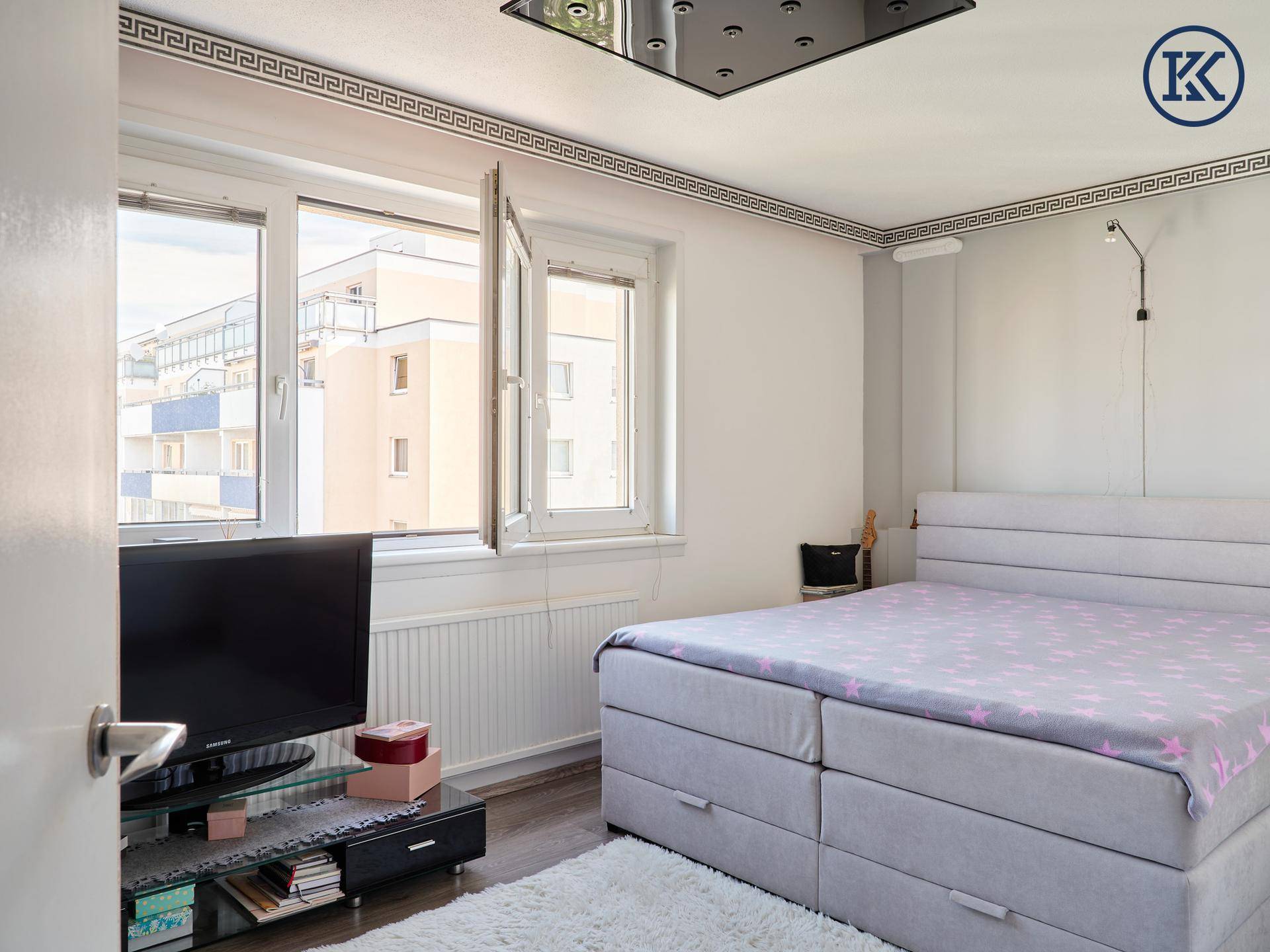 Helles Schlafzimmer mit großem Bett, Fernseher und einem Fenster mit Blick auf umliegende Gebäude.