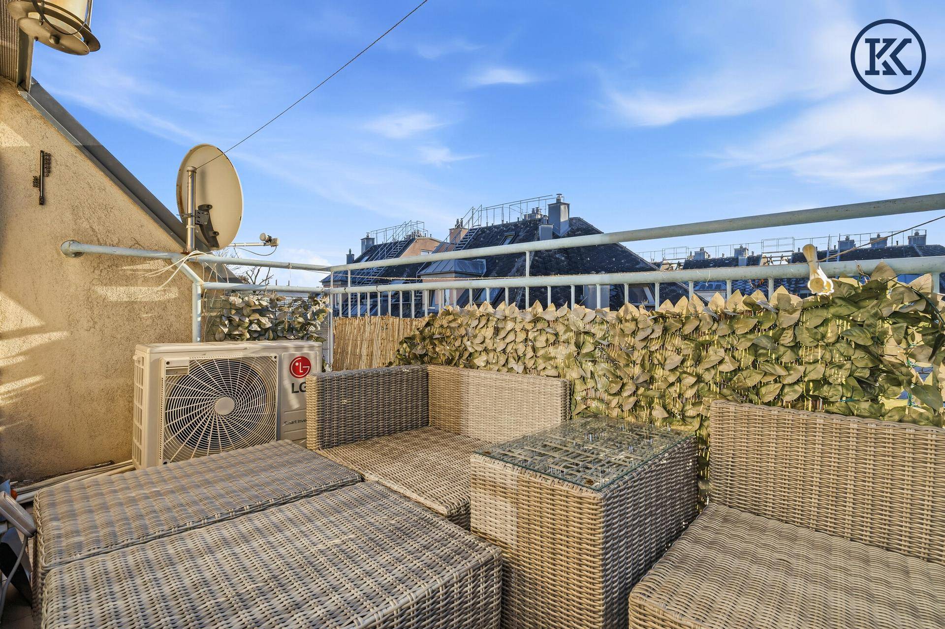 Sonniger Balkon mit Rattanmöbeln, Klimaanlage und Blick über die Dächer der Stadt.