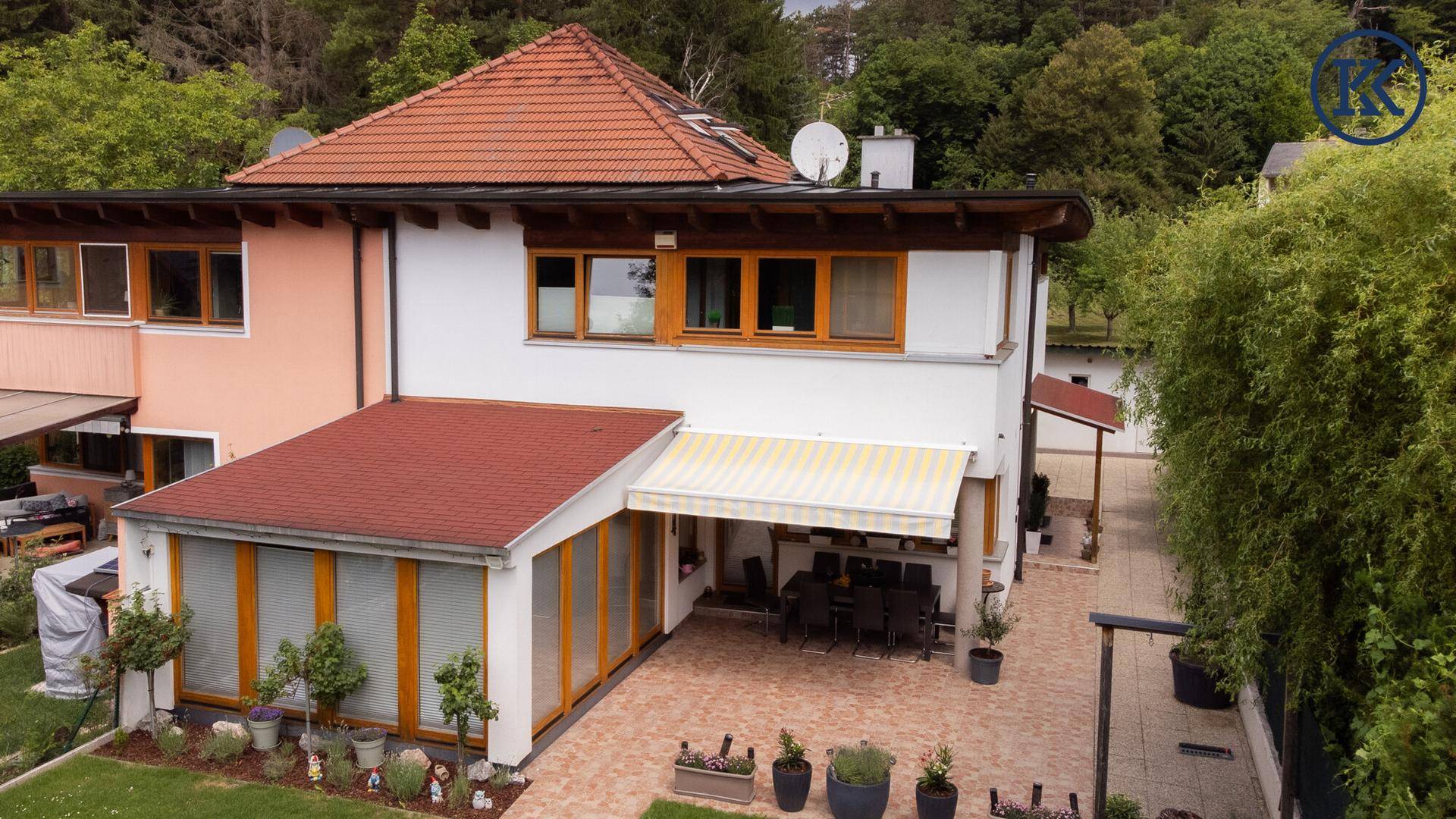 Rückansicht des Hauses mit großer Terrasse, Wintergarten und Markise, umgeben von Bäumen.