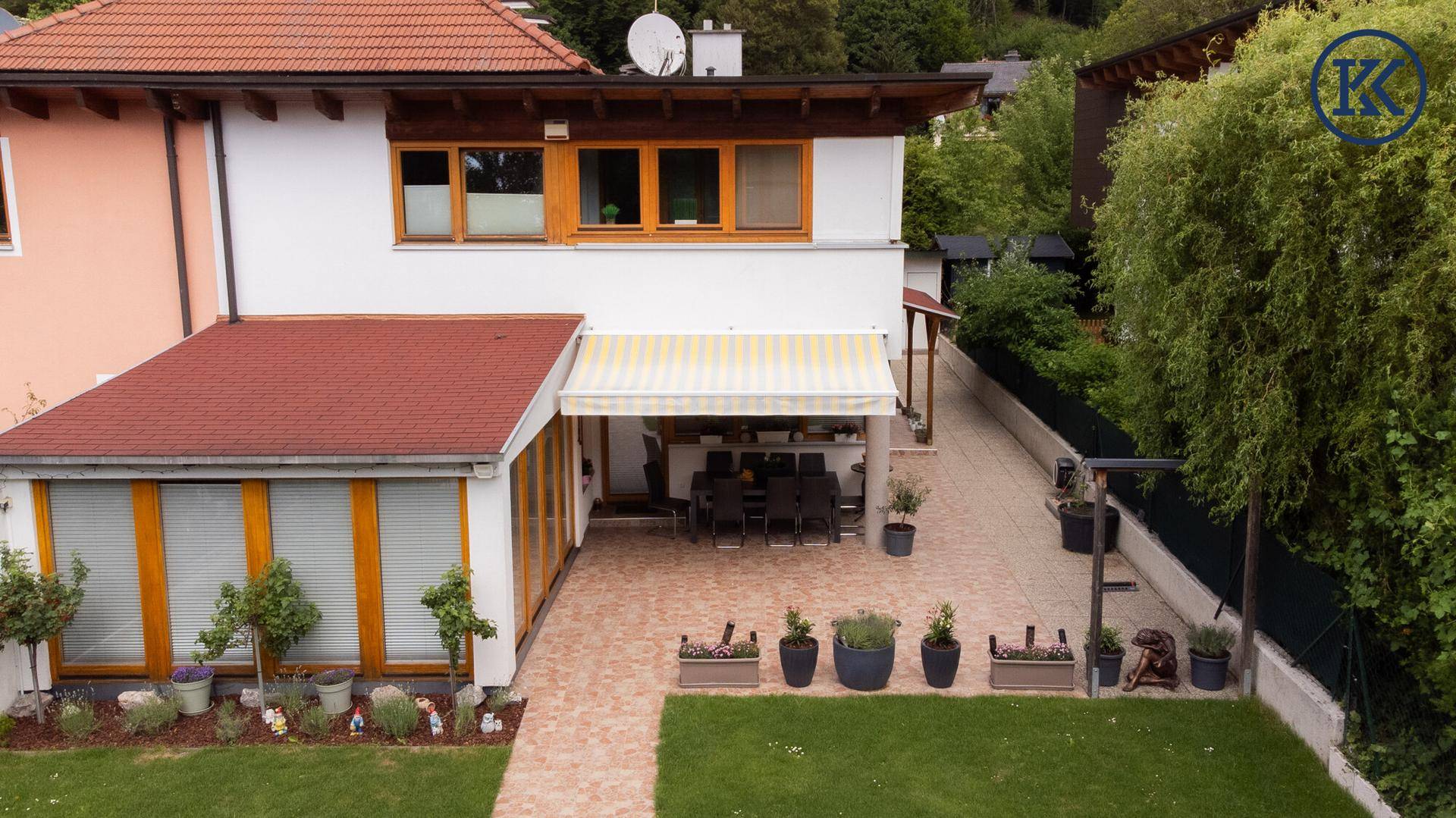 Ansicht der Hausrückseite mit großer gepflasterter Terrasse, Markise und angrenzendem Gartenbereich.