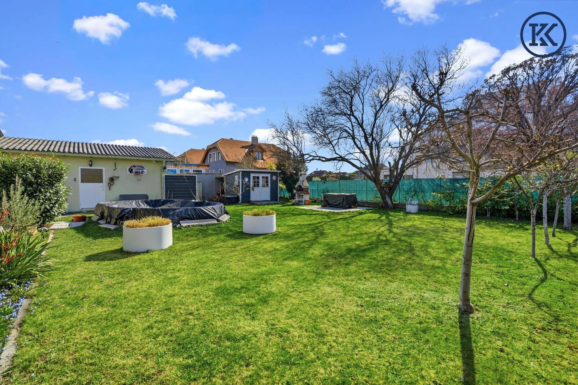 Geräumiger Garten mit Rasen, Gartenhaus und abgedecktem Poolbereich unter blauem Himmel.