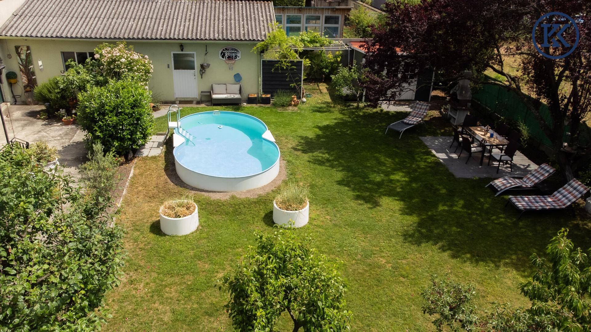 Gartenbereich mit Pool, schattigem Essplatz unter einem Baum und einem kleinen Gartenhaus.