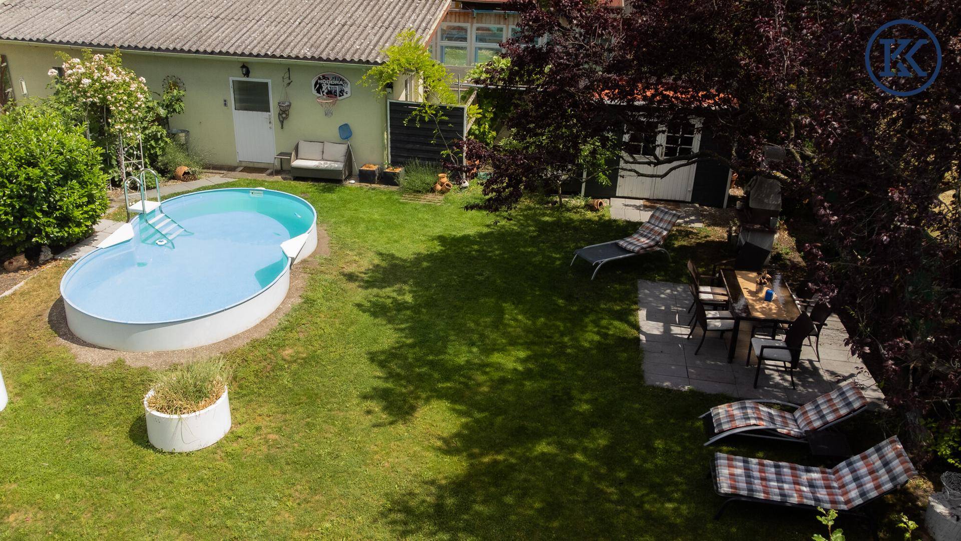 Gartenansicht mit Swimmingpool, Essbereich im Freien unter einem Baum und einem Gartenhaus.