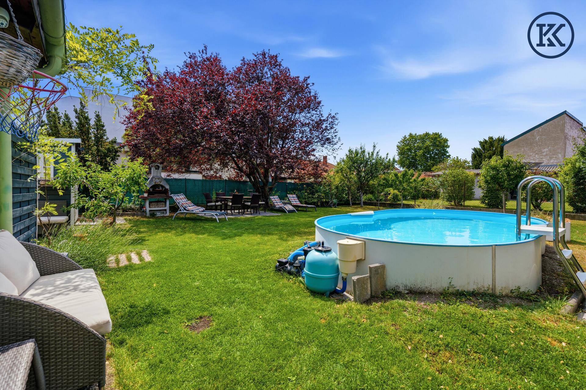 Blick in den Garten mit Pool, Grillplatz, Sonnenliegen und einem großen, schattenspendenden Baum.