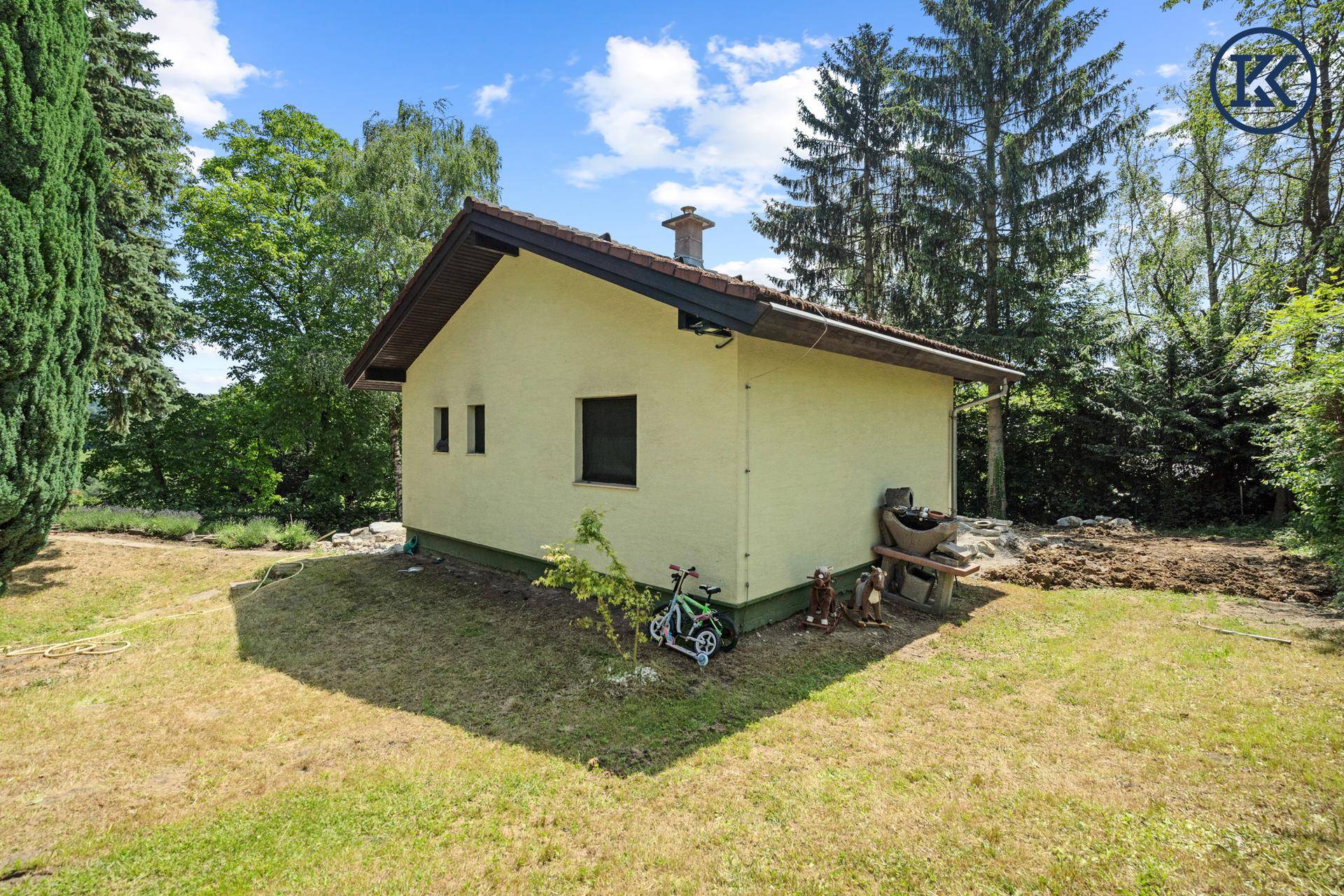 Kleineres Nebengebäude im Gartenbereich, umgeben von Rasen und hohen Bäumen unter blauem Himmel.