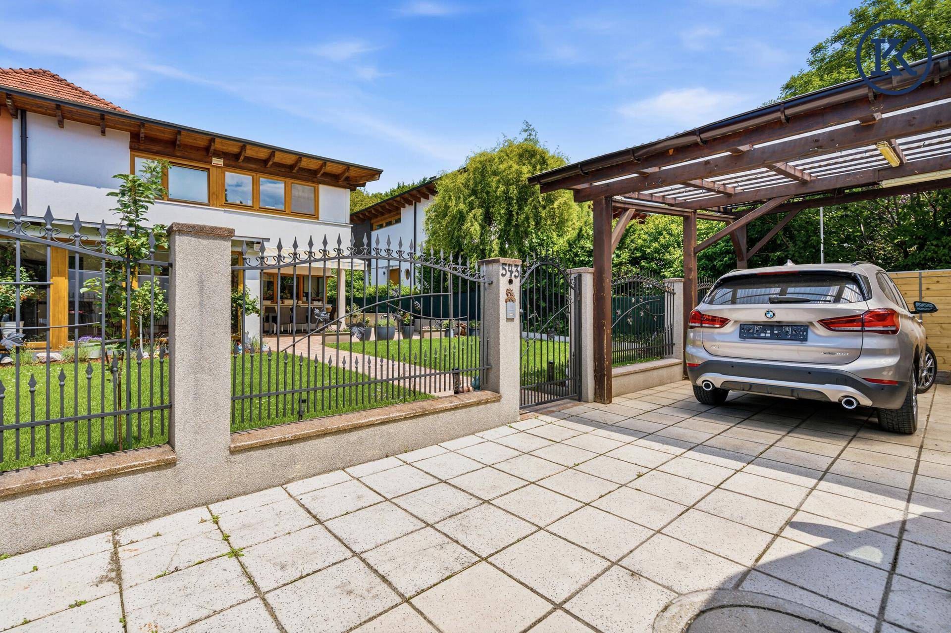 Eingangsbereich mit schmiedeeisernem Tor, gepflasterter Auffahrt und einem überdachten Carport.