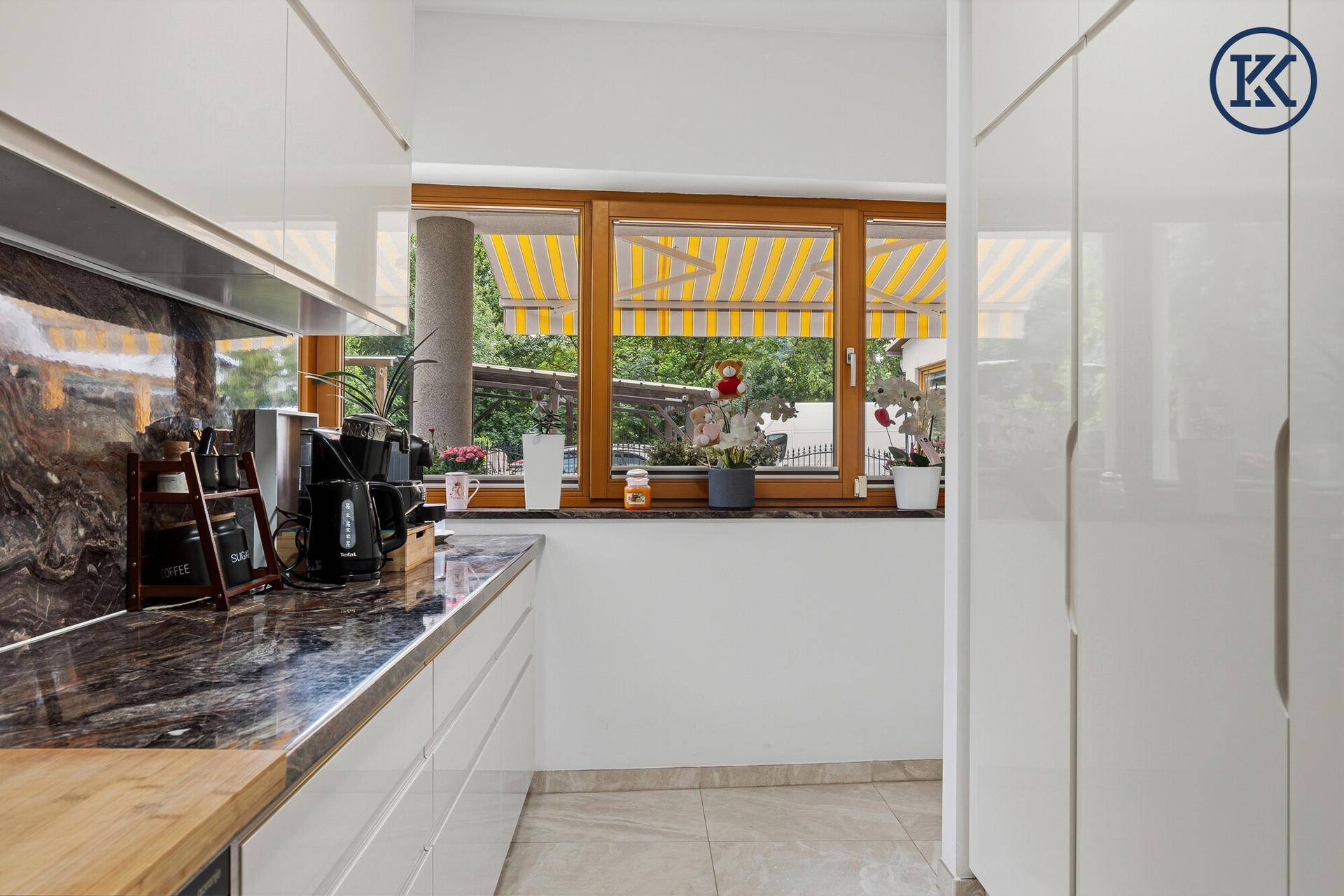 Helle Küche mit Fenster, Blick ins Grüne und weißen Schränken sowie dunkler Arbeitsplatte.