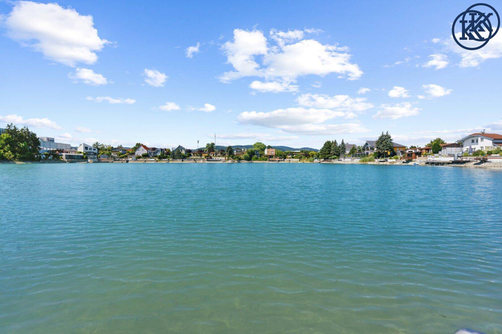 Panoramablick über den See mit Wohnhäusern entlang beider Uferseiten unter blauem Himmel.