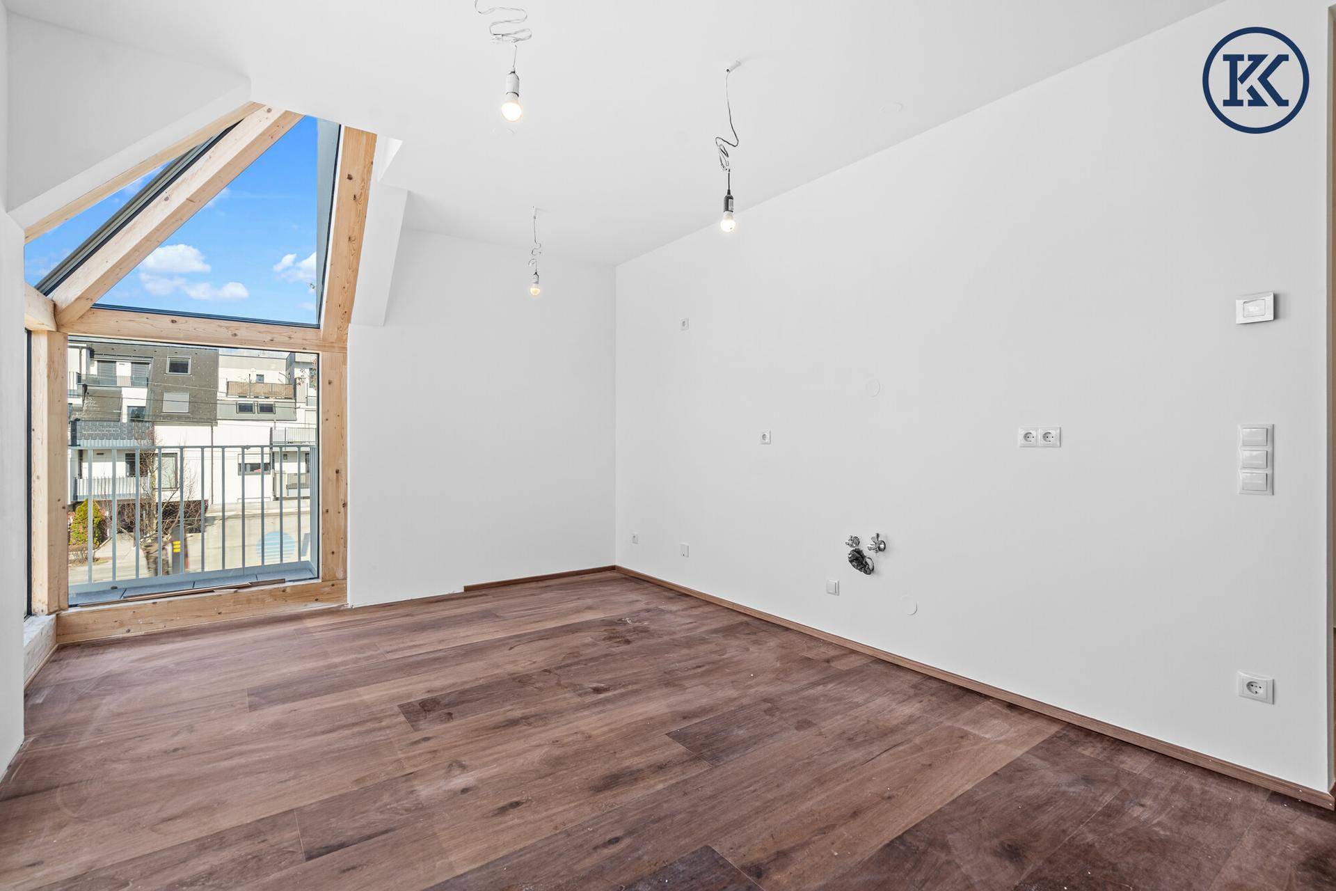 Helles Zimmer mit dunklem Holzboden, weißen Wänden und einem großen Giebelfenster.