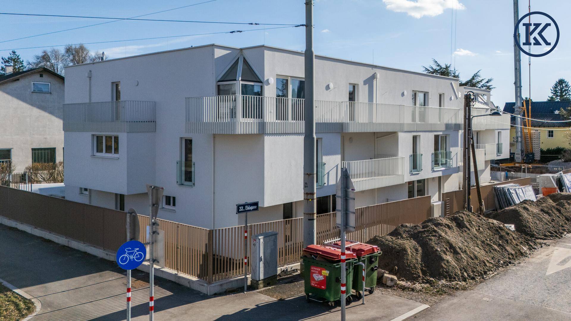 Eckansicht des Neubaus von der Straße, mit Bauabfällen und Mülltonnen im Vordergrund.