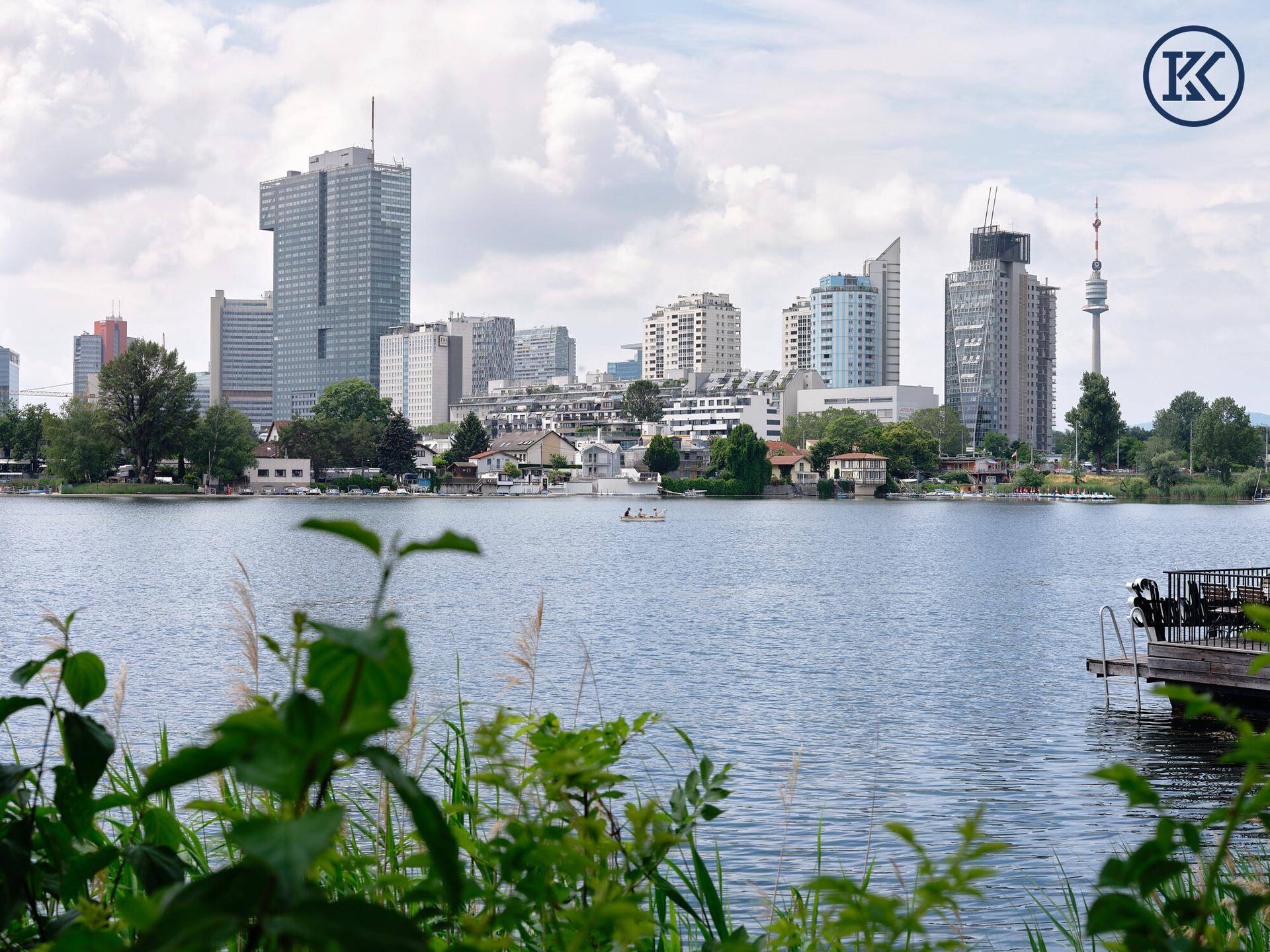 Weitläufiger Blick über den See zur modernen Wiener Skyline unter bewölktem Himmel.