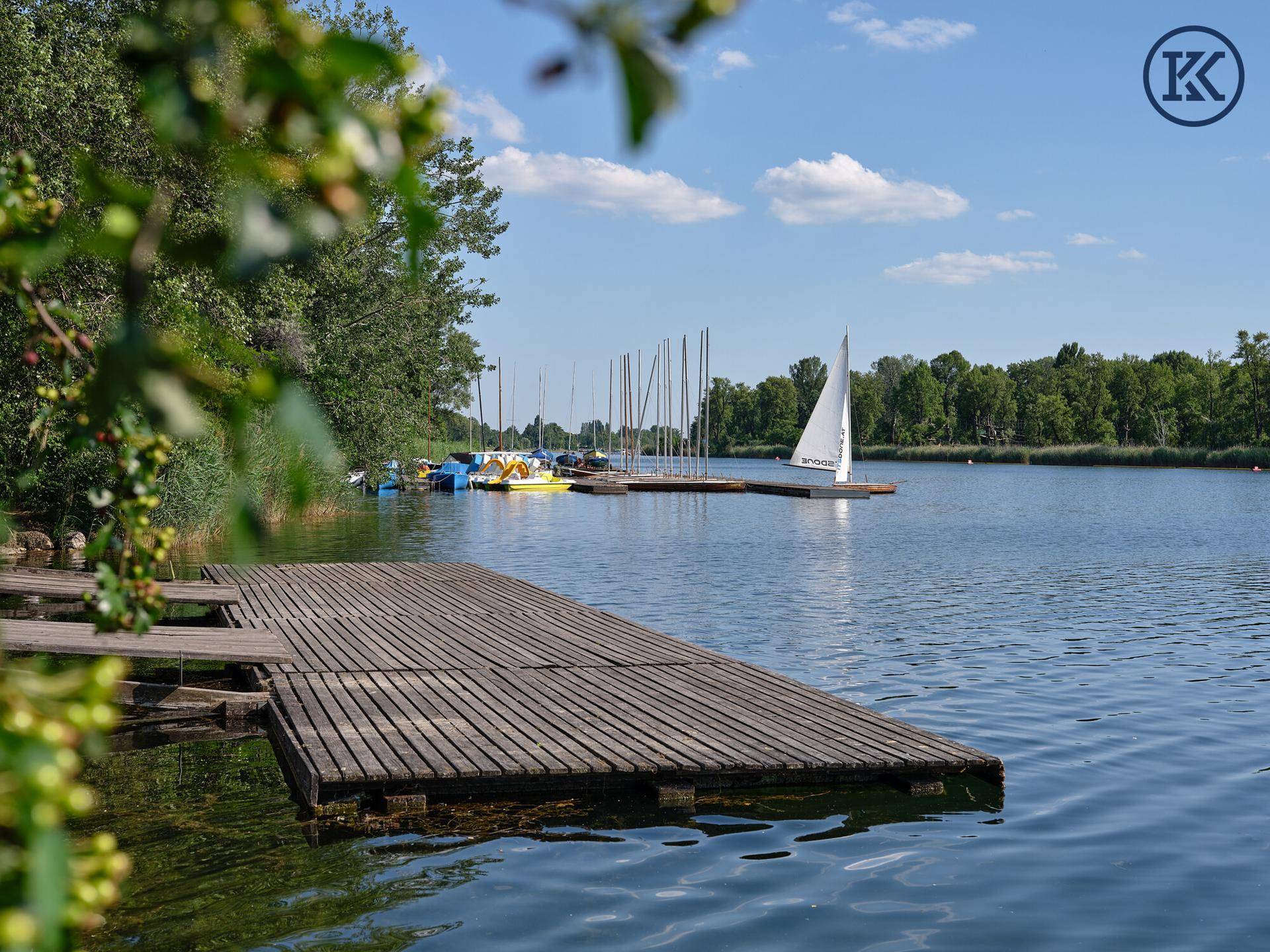 Holzsteg am See mit Segelbooten im Hintergrund und klarem blauen Himmel.