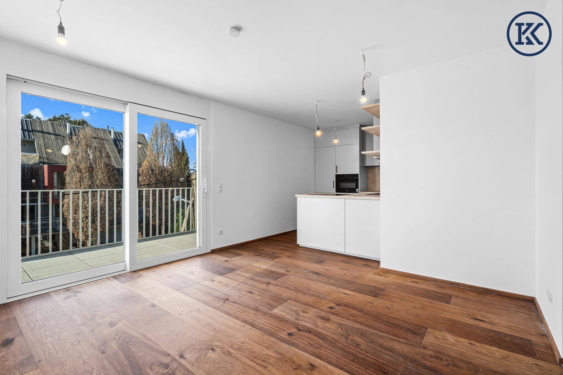 Heller Wohnbereich mit Holzboden, offener Küche und Zugang zu einem Balkon mit Blick ins Grüne.