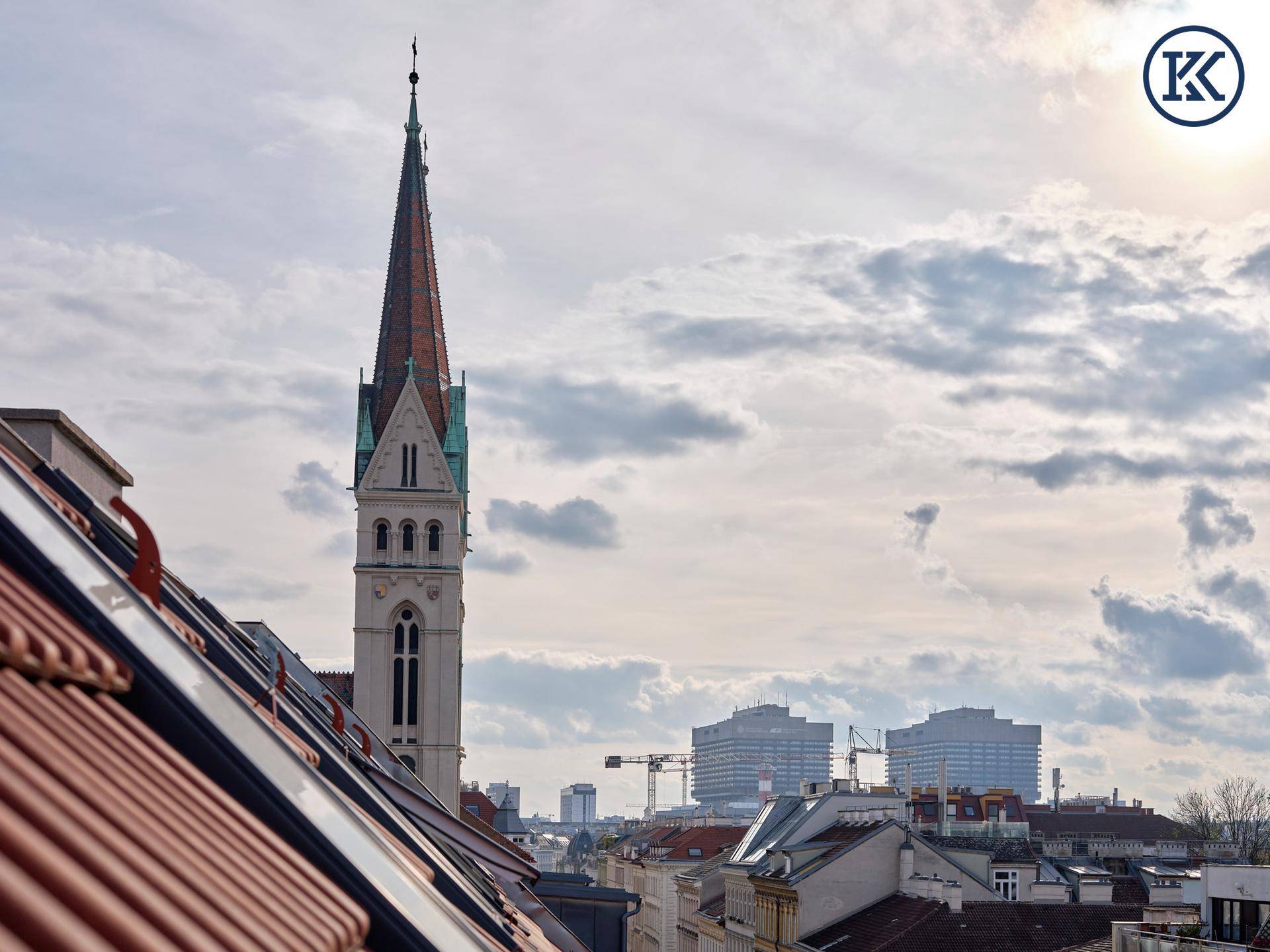 Blick auf eine imposante Kirchturmspitze über den Dächern der Stadt unter bewölktem Himmel.