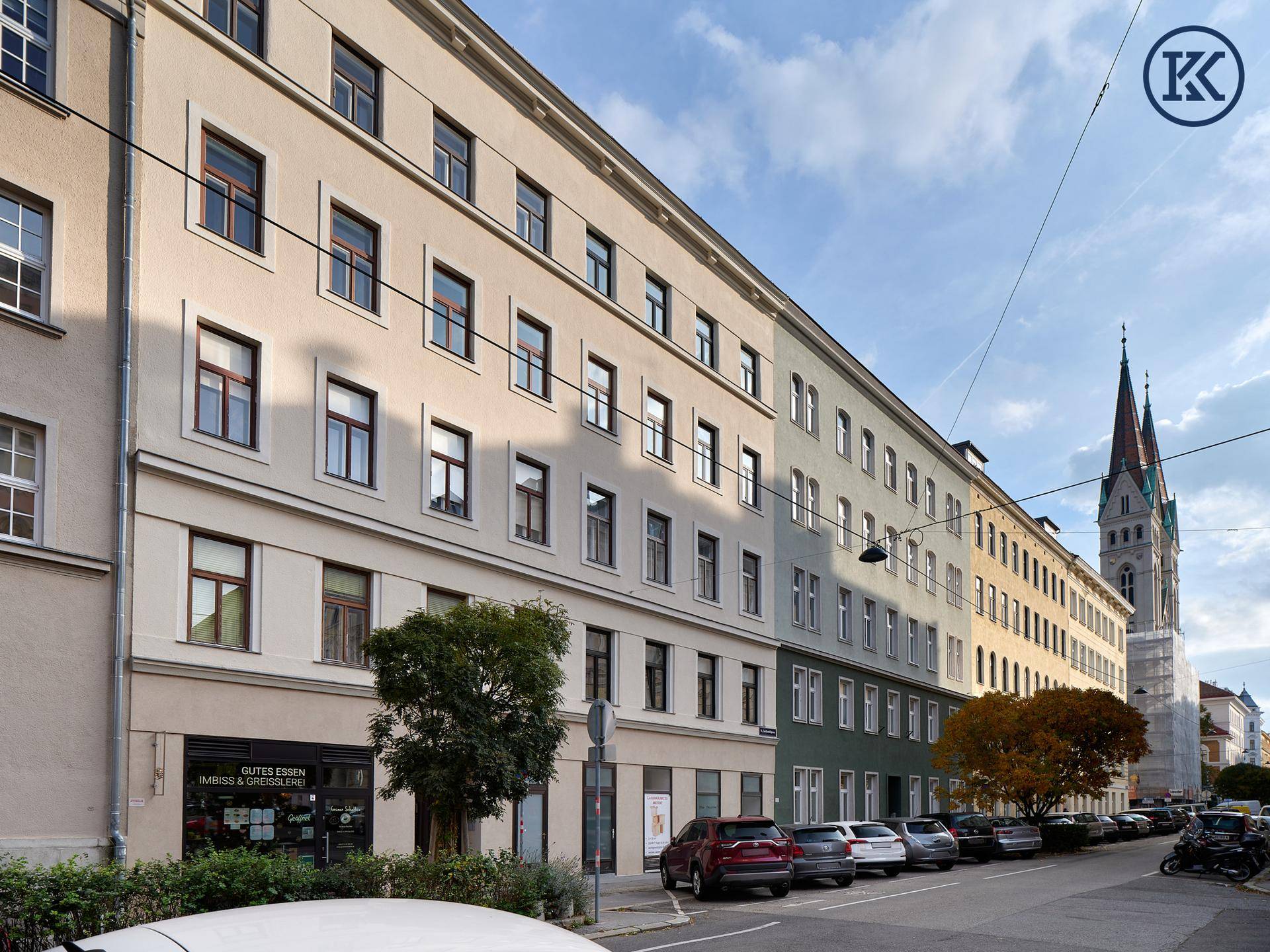 Straßenansicht mit einer Reihe von Gebäuden, parkenden Autos und einem Kirchturm im Hintergrund.