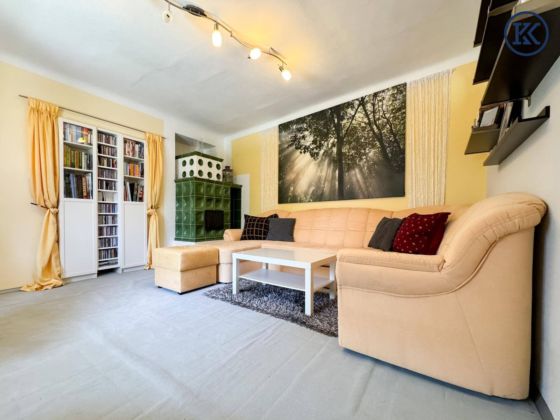 Geräumiges Wohnzimmer mit hellem Teppichboden, großem Sofa und grünem Kachelofen.