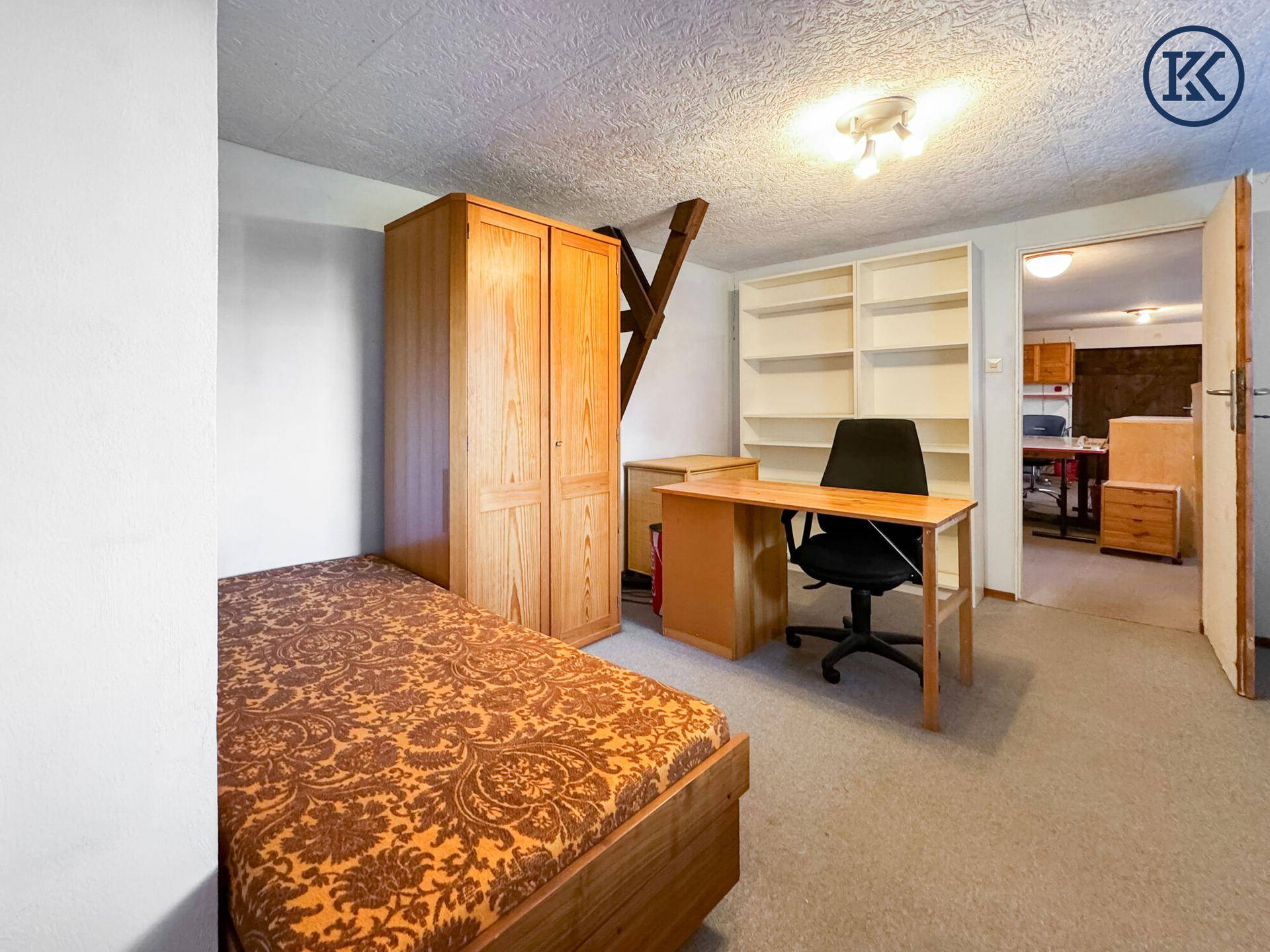 Schlafzimmer mit Einzelbett, Holzschrank und Schreibtisch, das in einen weiteren Raum blickt.