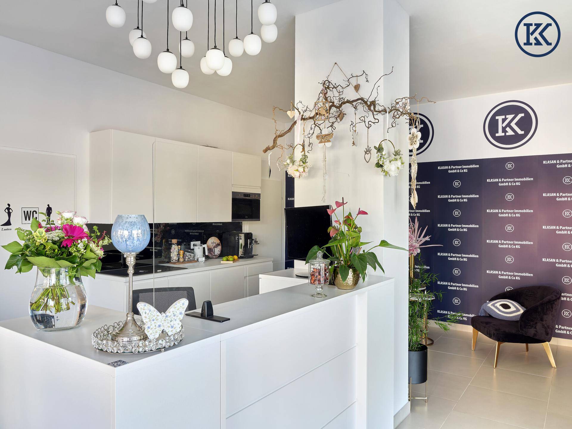 Empfangsbereich mit moderner Theke, Blumenarrangements und einer Küchenzeile im Hintergrund.