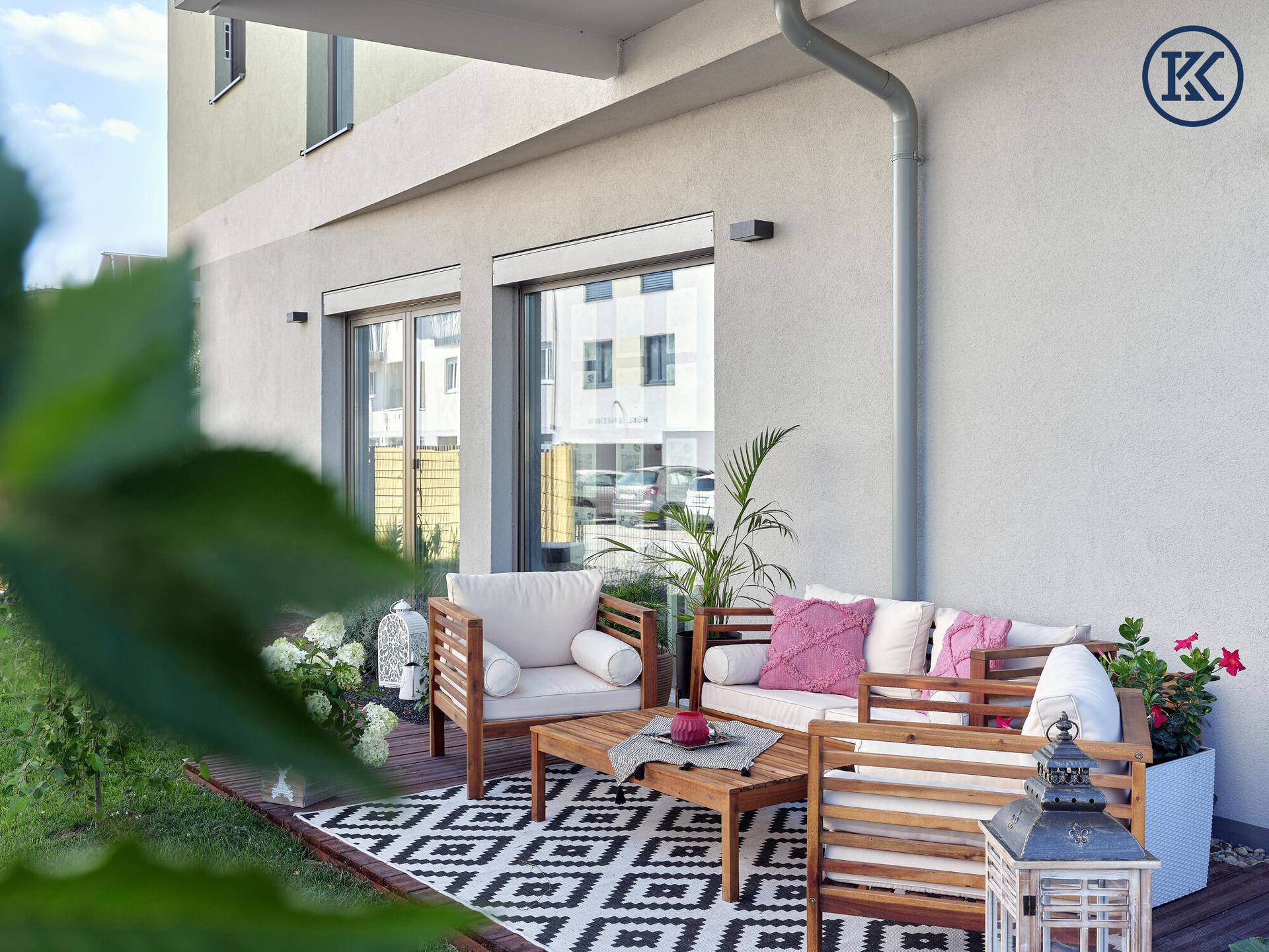 Gemütliche Terrasse mit Holzmöbeln, Kissen und Pflanzen, angrenzend an ein Gebäude.