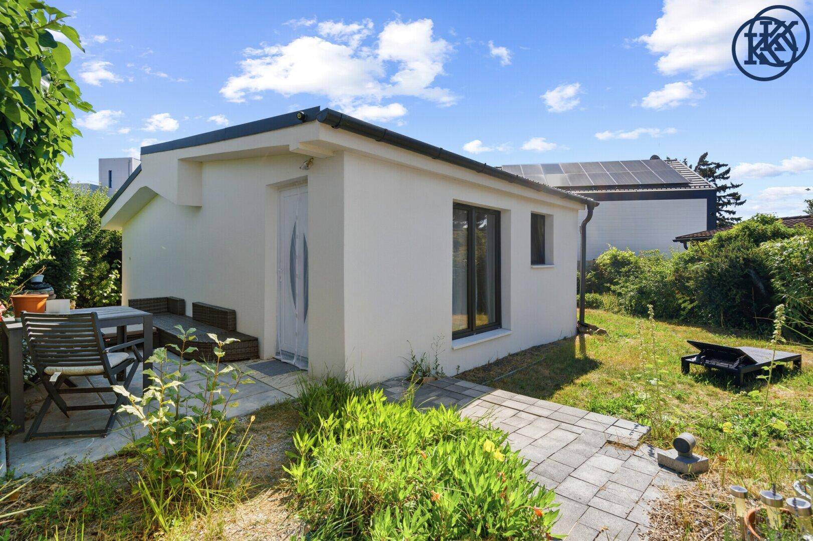 Weißes Gebäude mit gepflasterter Terrasse und Gartenmöbeln, umgeben von grüner Vegetation.