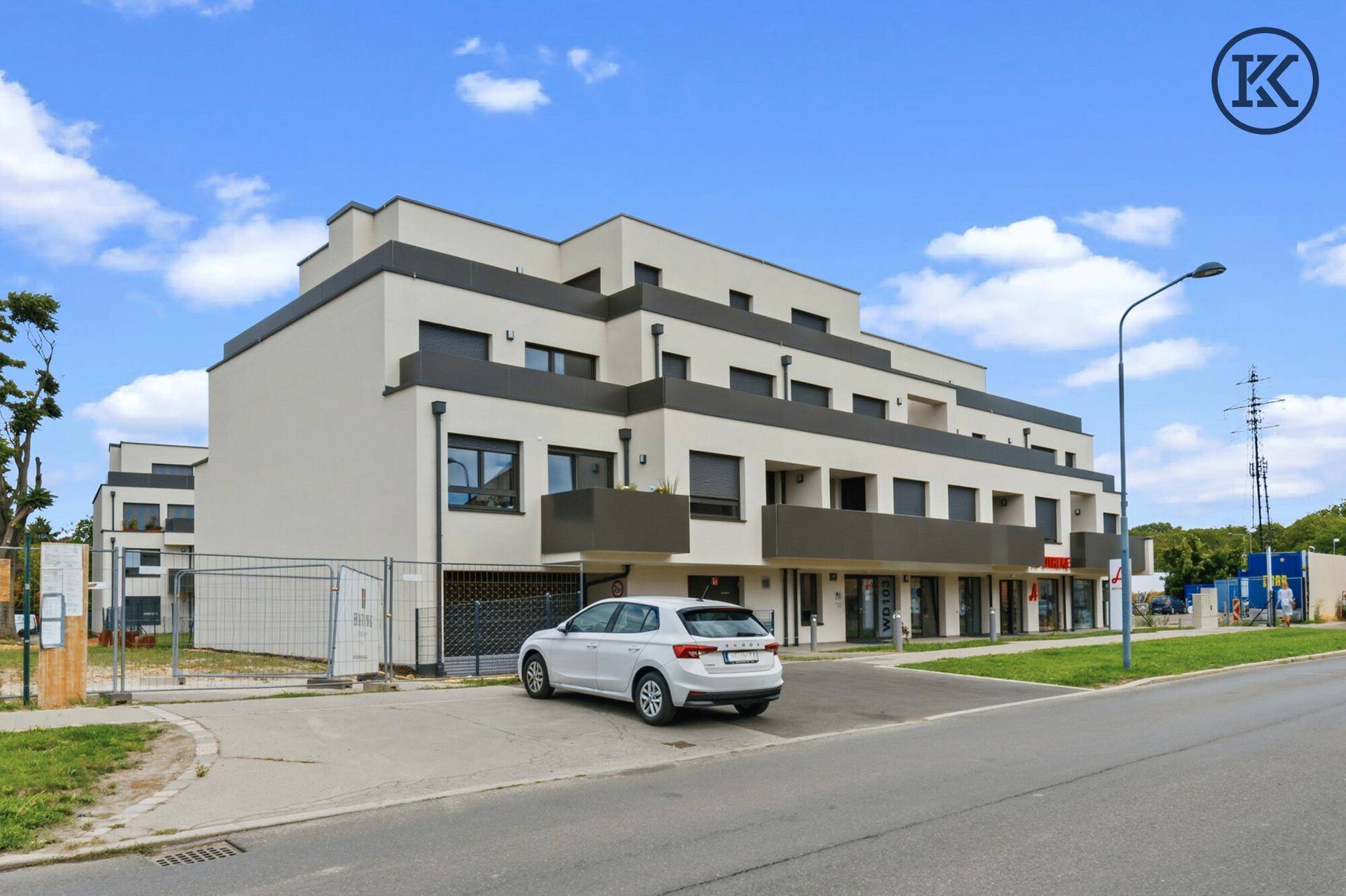 Modernes Wohngebäude mit Parkplatz und Geschäften im Erdgeschoss unter blauem Himmel.