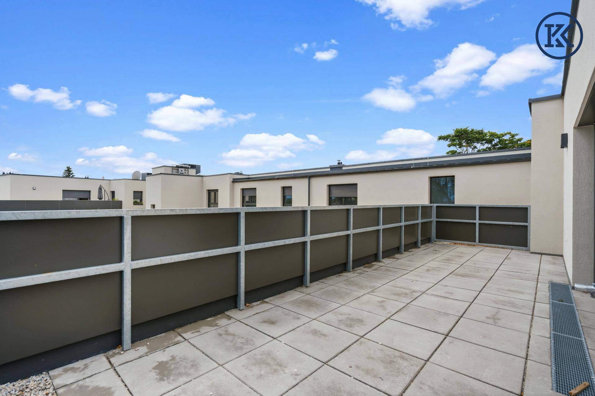 Geräumige Dachterrasse mit Fliesenboden und moderner Brüstung unter blauem Himmel.