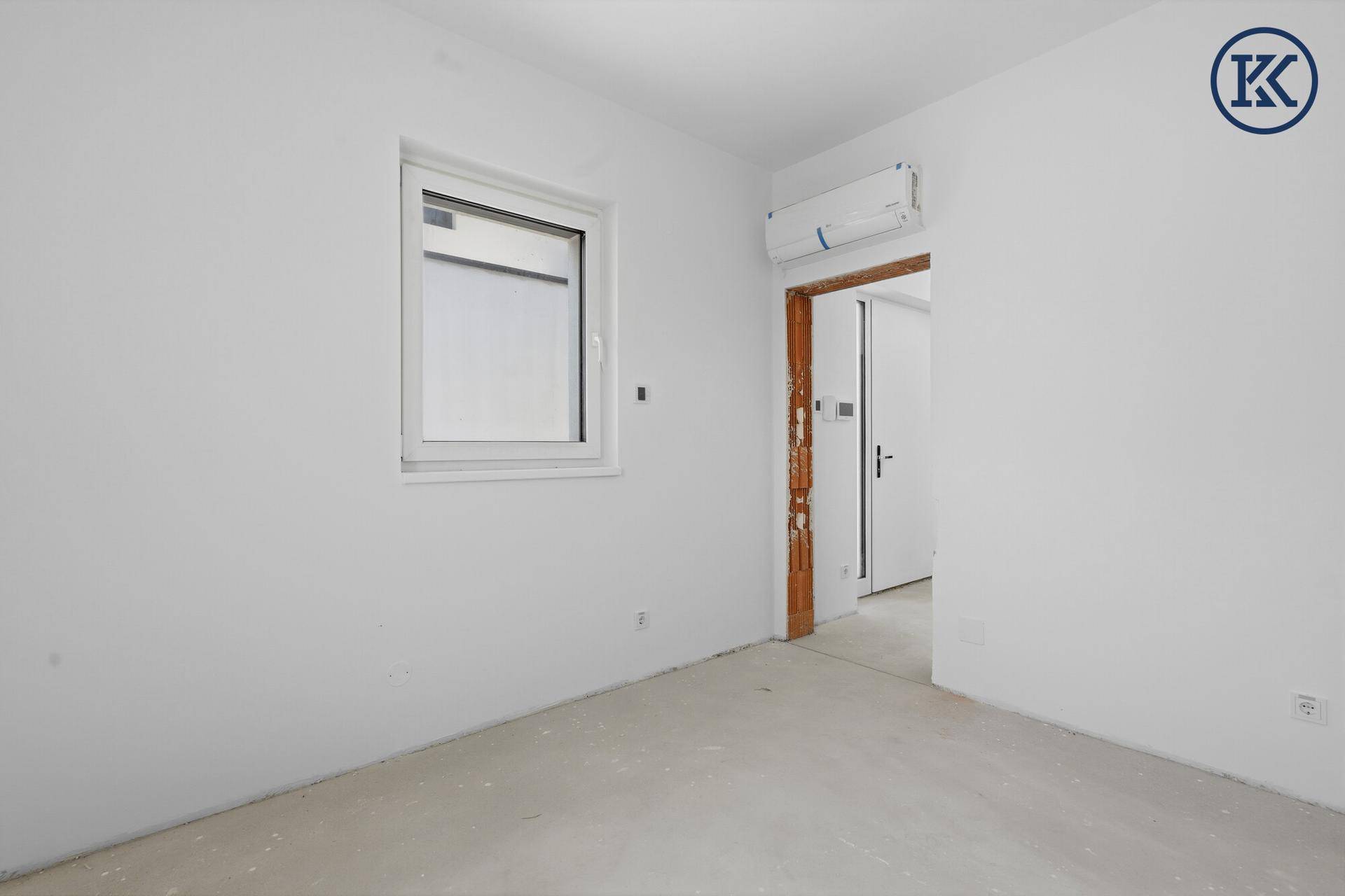 Leerer Raum im Neubau mit weiß gestrichenen Wänden, einem Fenster und vorbereiteter Klimaanlage.