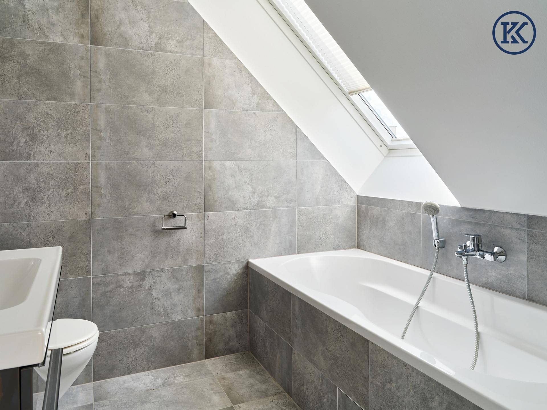 Helles Badezimmer mit grauen Fliesen, Badewanne und Waschtisch unter einem schrägen Dachfenster.