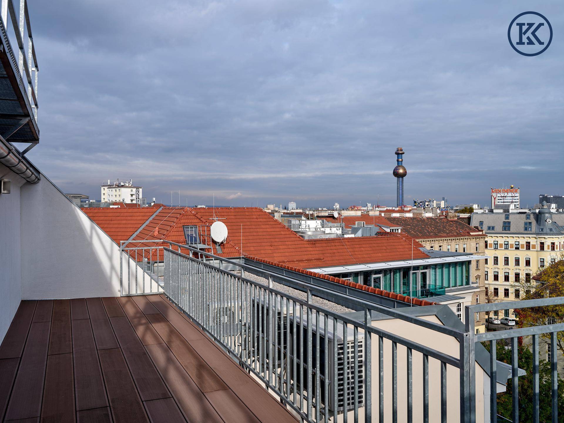 Dachterrasse mit Holzboden, Metallgeländer und weitem Blick über die Dächer der Stadt und einen Turm.