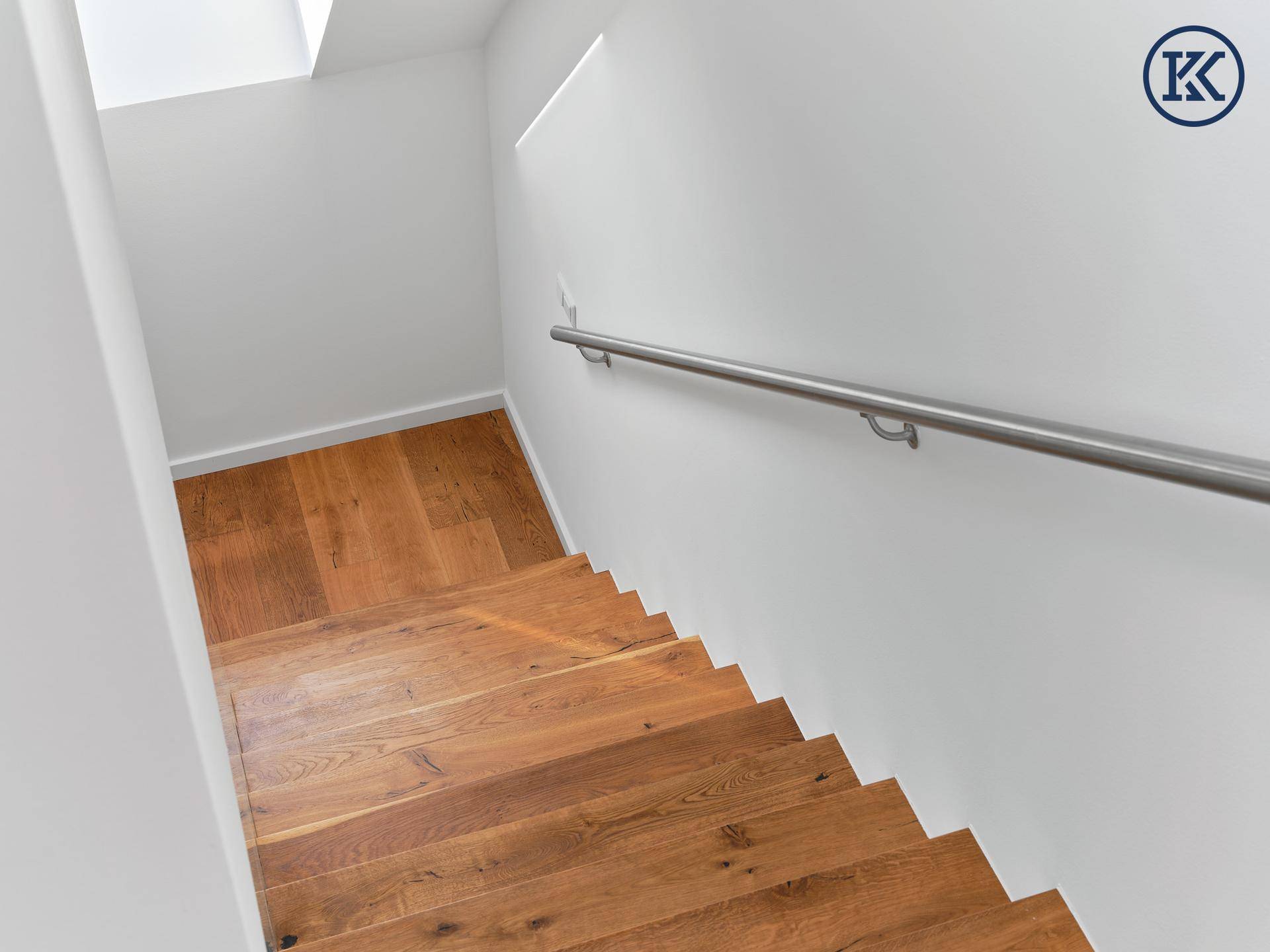 Treppenabgang mit Holzdielen und schlichtem Edelstahlhandlauf an der weißen Wand.