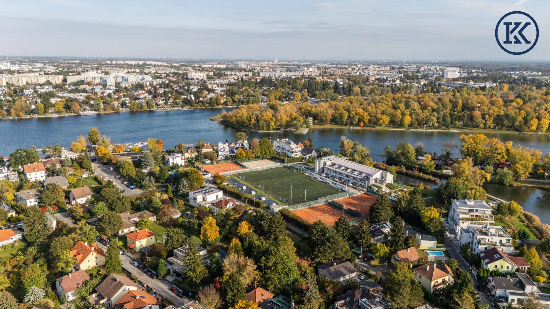 Luftaufnahme einer Wohngegend mit Seen, Tennisplätzen und einem Fußballfeld, umgeben von herbstlichen Bäumen.