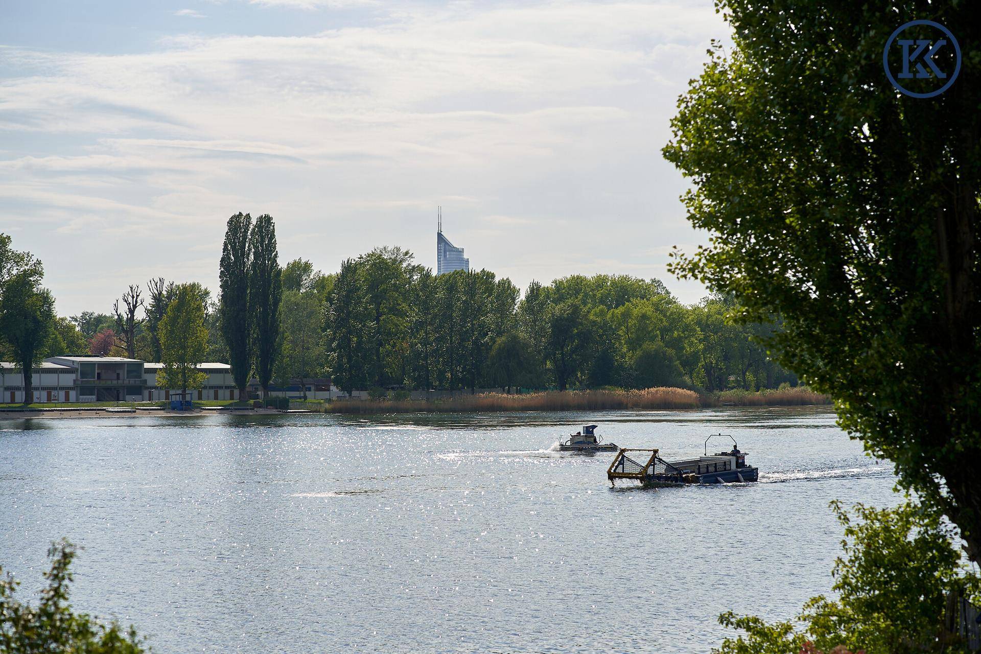 Malerischer Blick auf den See mit vorbeifahrenden Booten und grünen Ufern unter blauem Himmel.