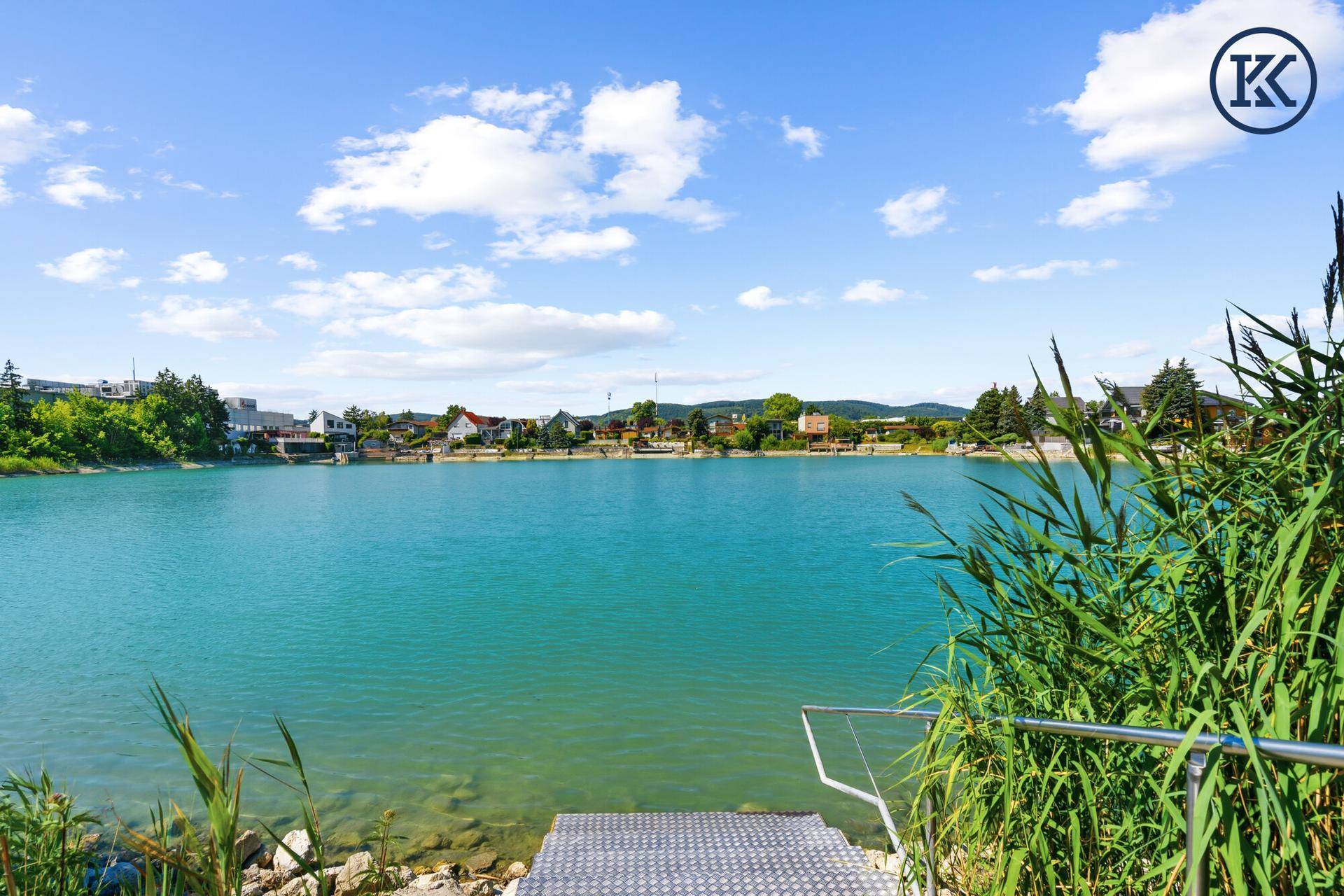 Malerischer Seeblick mit Schilf im Vordergrund und Häusern am Ufer unter blauem Himmel.