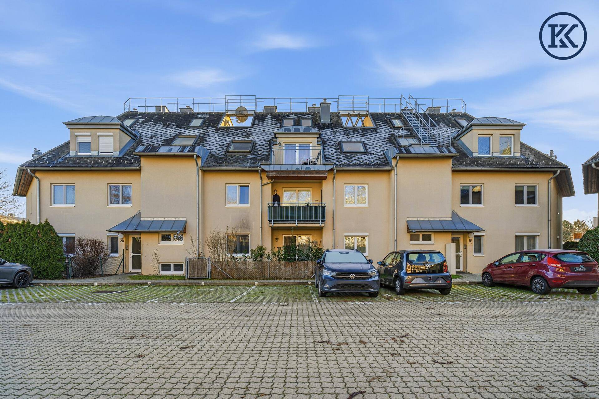 Mehrfamilienhaus mit Mansarddach, Balkonen und Außenparkplätzen unter blauem Himmel.