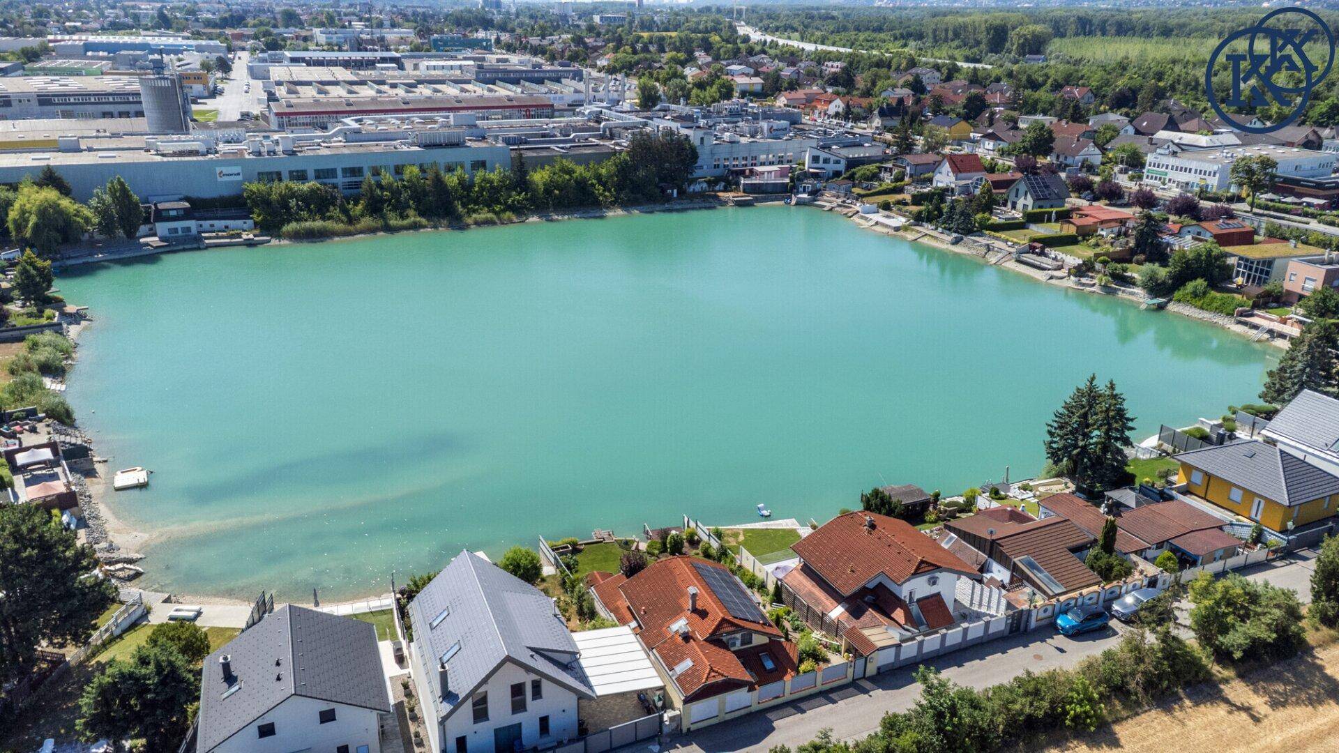 Weitläufige Luftaufnahme des Sees, umgeben von Wohngebieten und Industrieanlagen unter blauem Himmel.