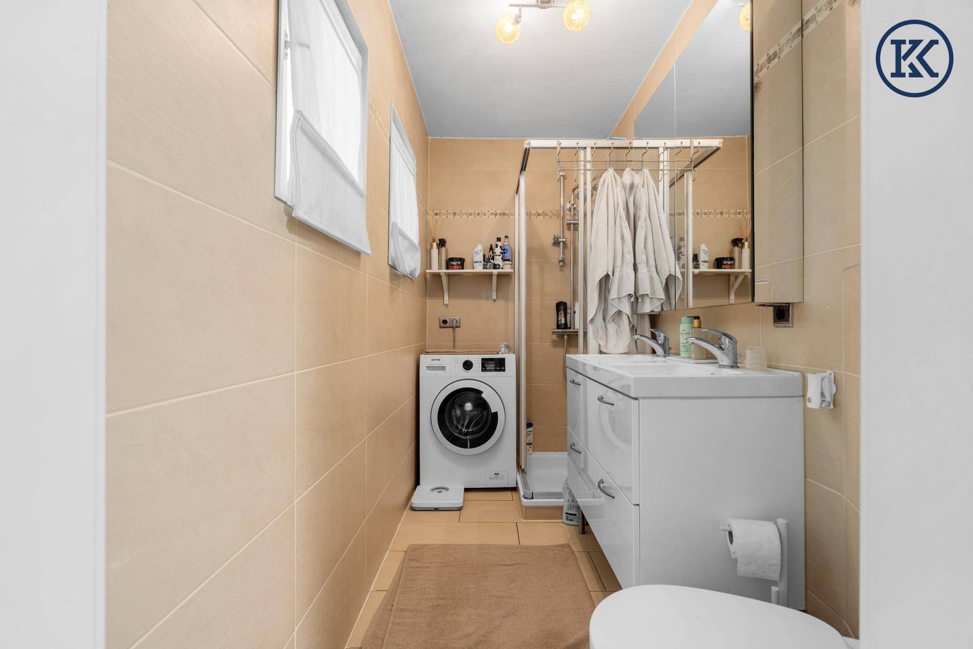 Badezimmer mit beigen Fliesen, Dusche, Waschbecken und integrierter Waschmaschine.