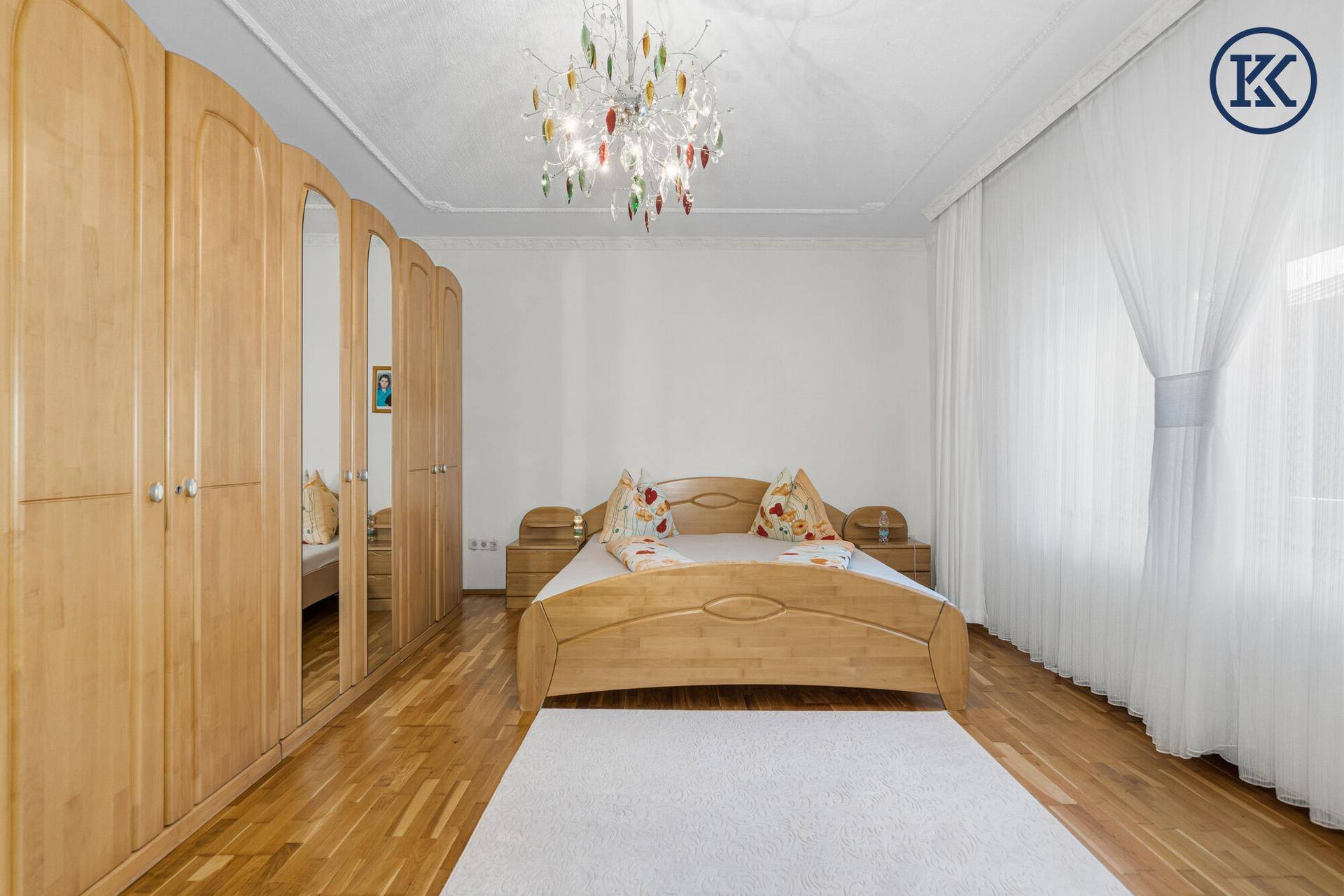 Helles Schlafzimmer mit Holzmöbeln, Parkettboden und großem Fenster mit Vorhängen.