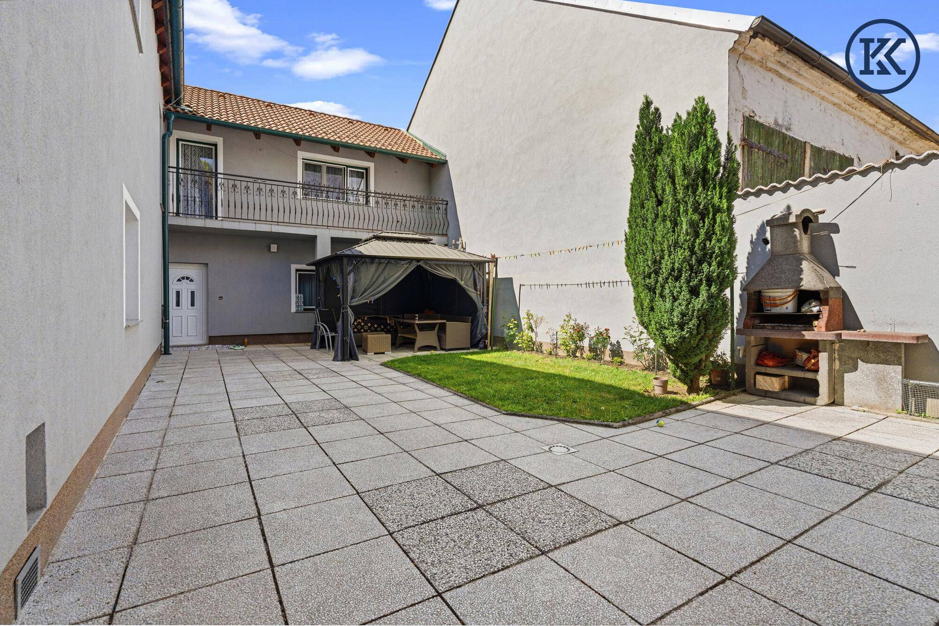 Gepflasterter Innenhof mit Rasenfläche, Grillplatz und überdachtem Sitzbereich im Freien.
