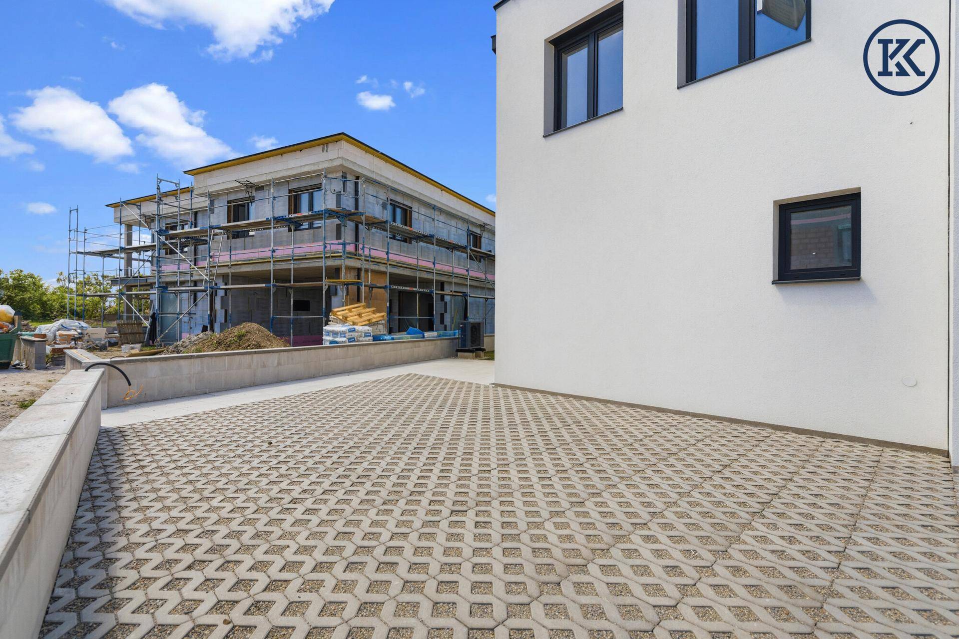 Außenansicht eines Neubaus mit gepflastertem Bereich und einem weiteren Gebäude im Bau im Hintergrund.