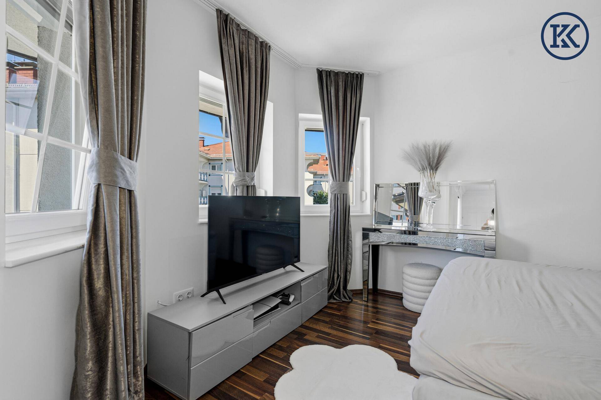 Schlafzimmer mit Fernseher, dunklem Holzboden und Fenstern, die viel Tageslicht hereinlassen.