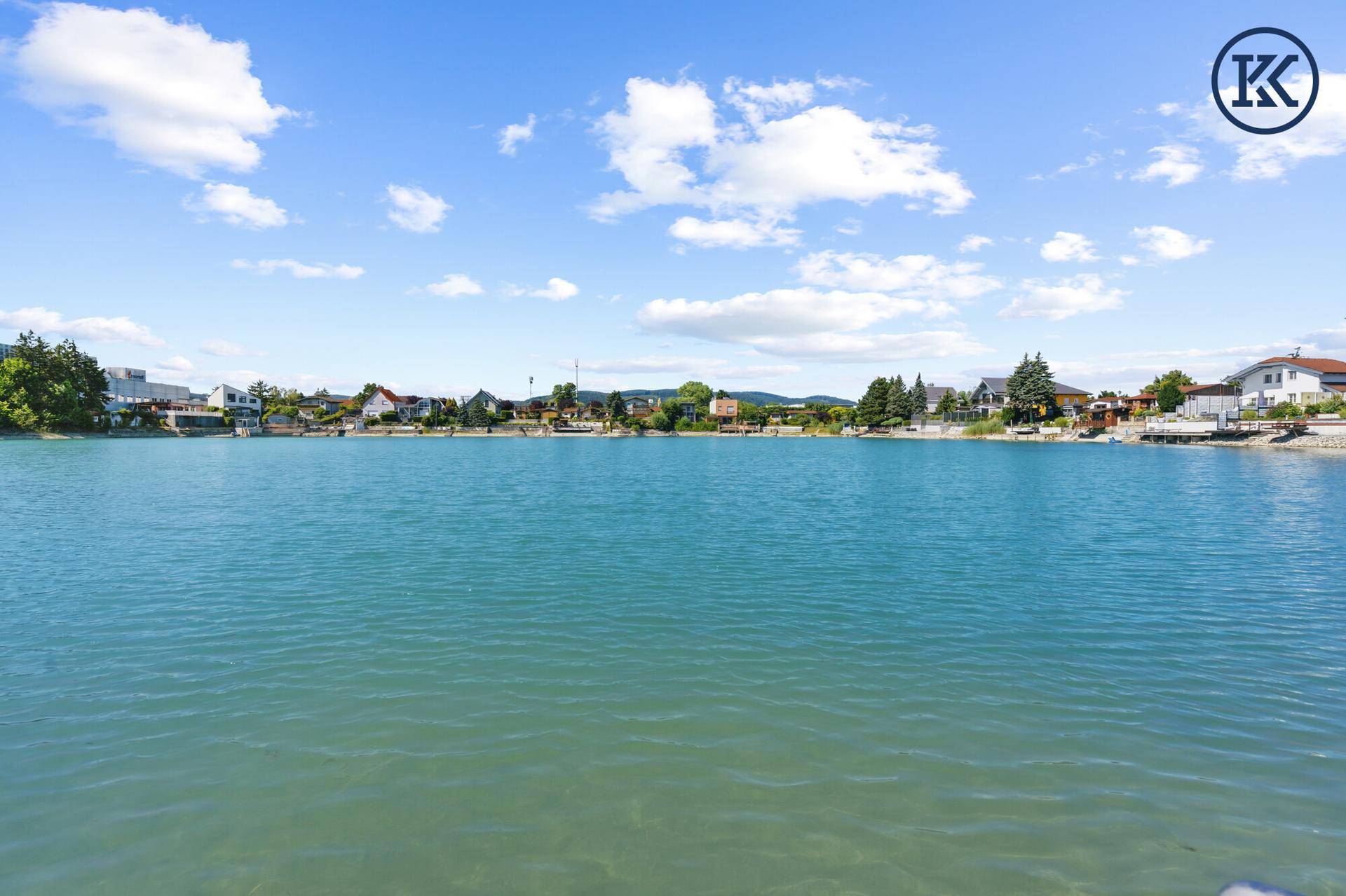 Panoramablick über den See mit Wohnbebauung am gegenüberliegenden Ufer unter blauem Himmel.