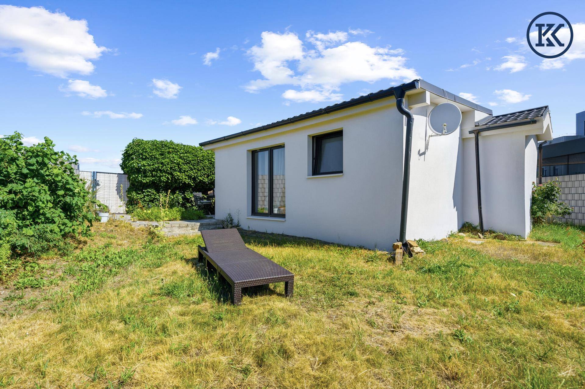 Außenansicht des kleinen weißen Hauses mit sonnigem Garten und Liegestuhl.