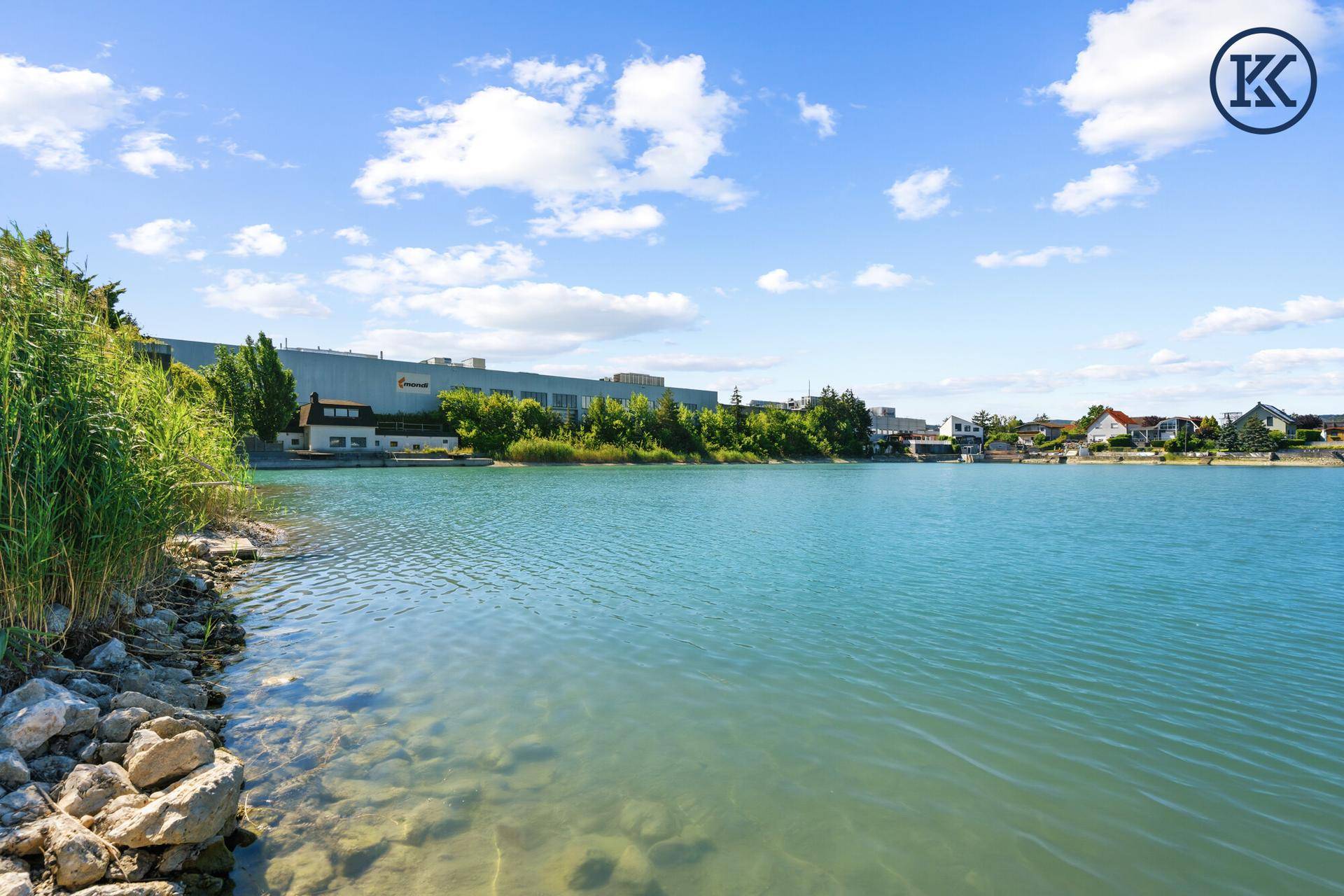 Weitläufiger Blick über den See mit Industriegebäude und grüner Ufervegetation im Hintergrund.
