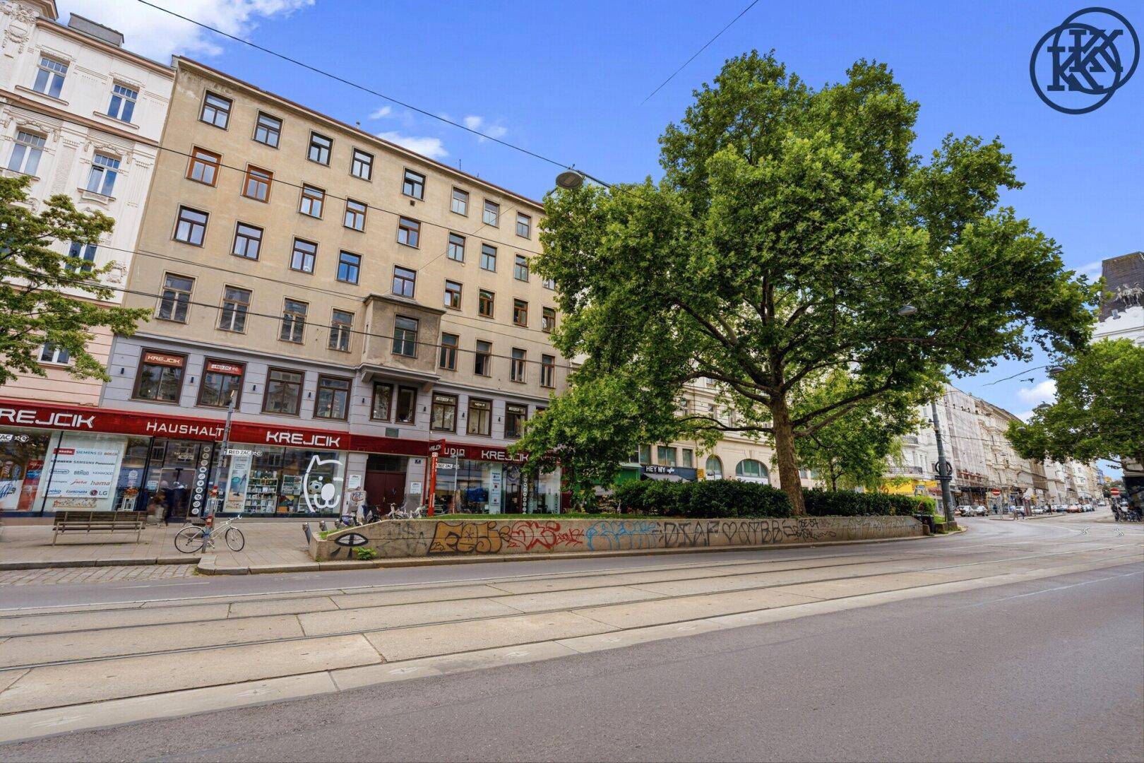 Gebäudeansicht mit Geschäften im Erdgeschoss, Bäumen und einer breiten Straße.