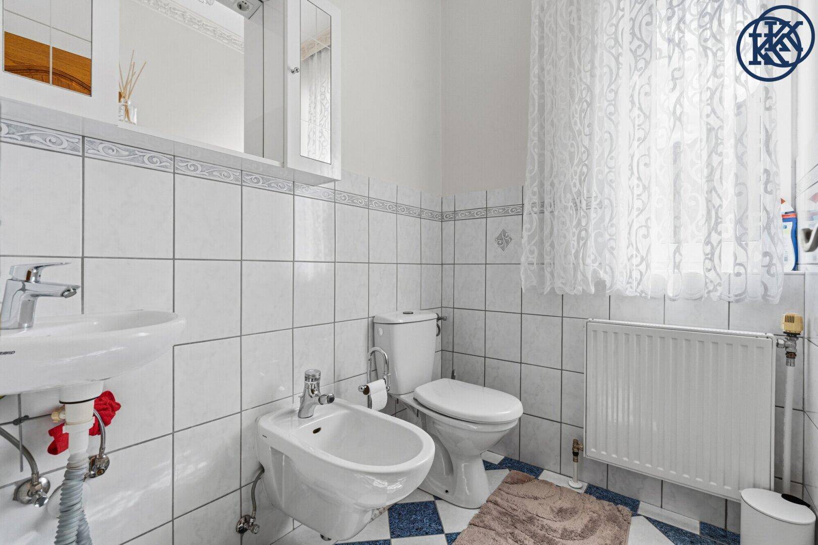 Badezimmer mit weißen und blauen Fliesen, Waschbecken, Toilette, Bidet und Fenster.