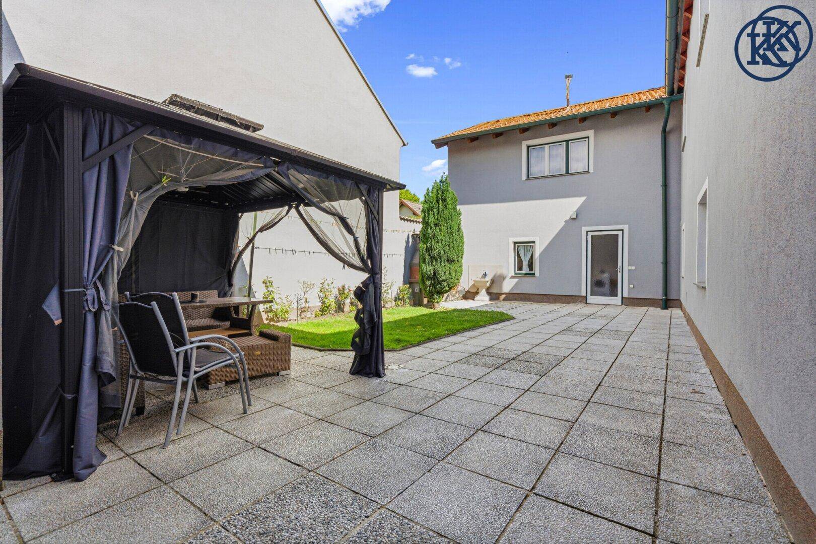 Weitläufige gepflasterte Terrasse mit Rasenfläche und Gartenlaube, umgeben von Gebäuden.