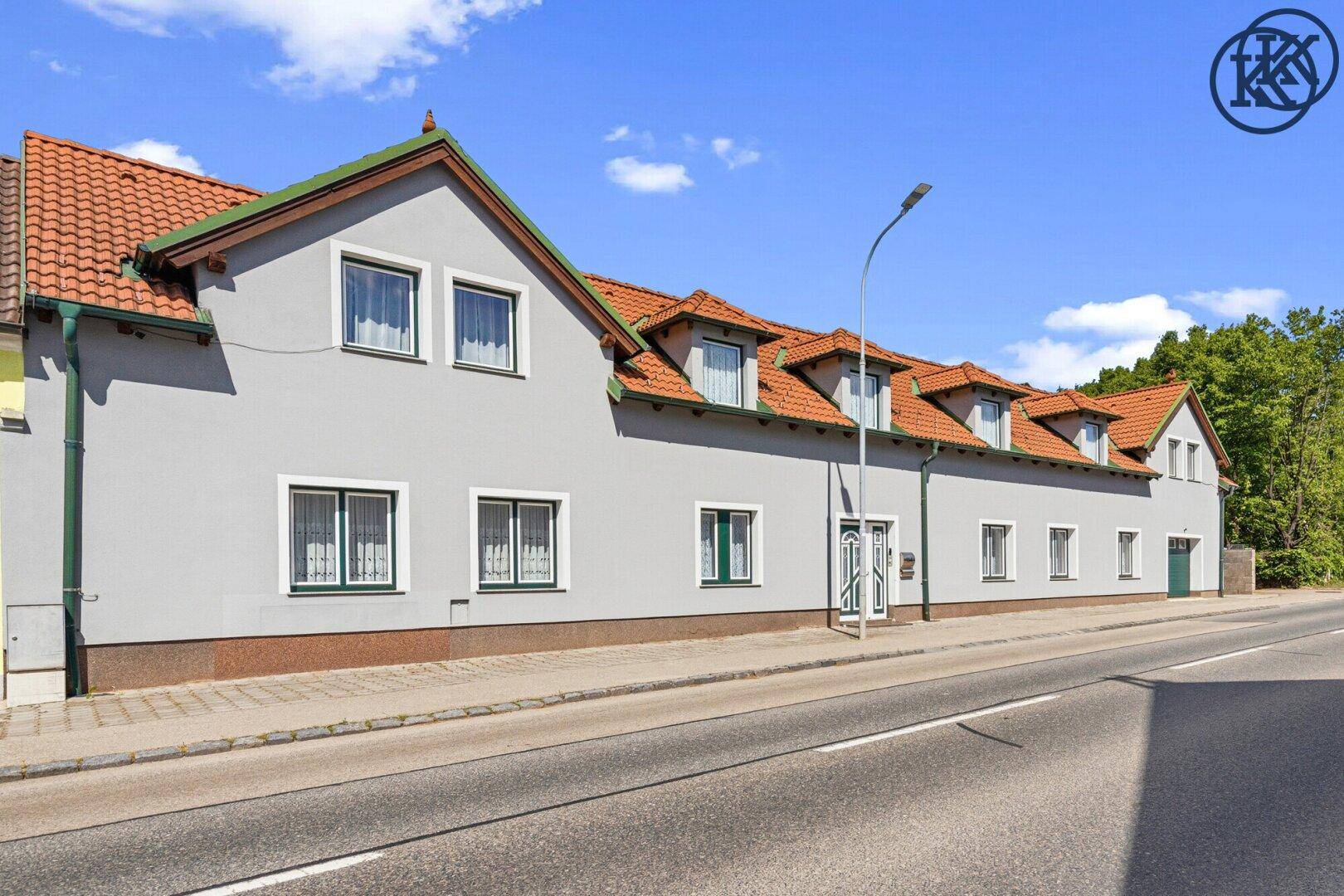 Lange Hausfassade mit mehreren Fenstern, Gauben und einem traditionellen Ziegeldach an einer Straße.