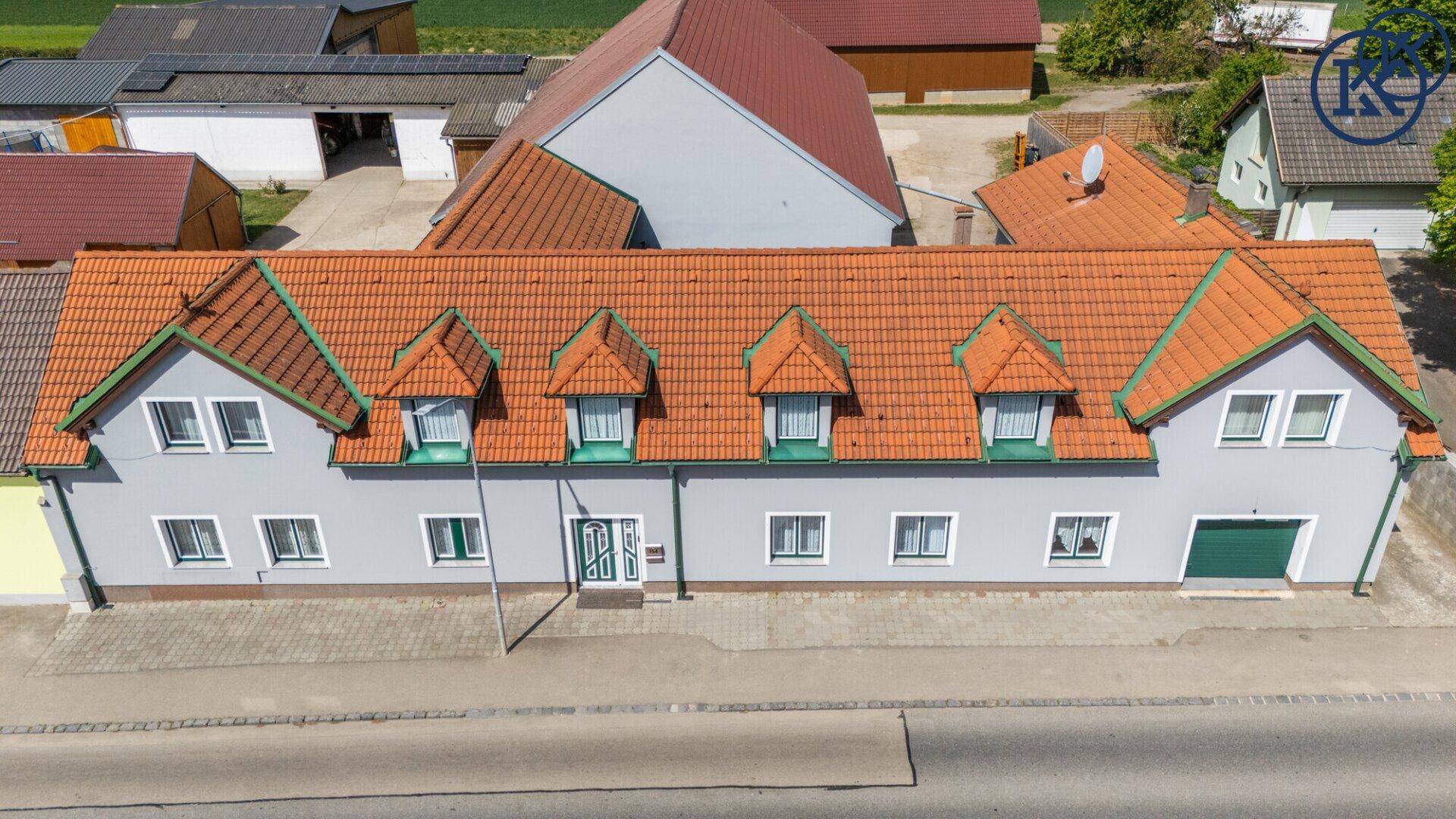 Frontale Luftaufnahme des langen Gebäudes mit Ziegeldach und Gauben, umgeben von Grünflächen.