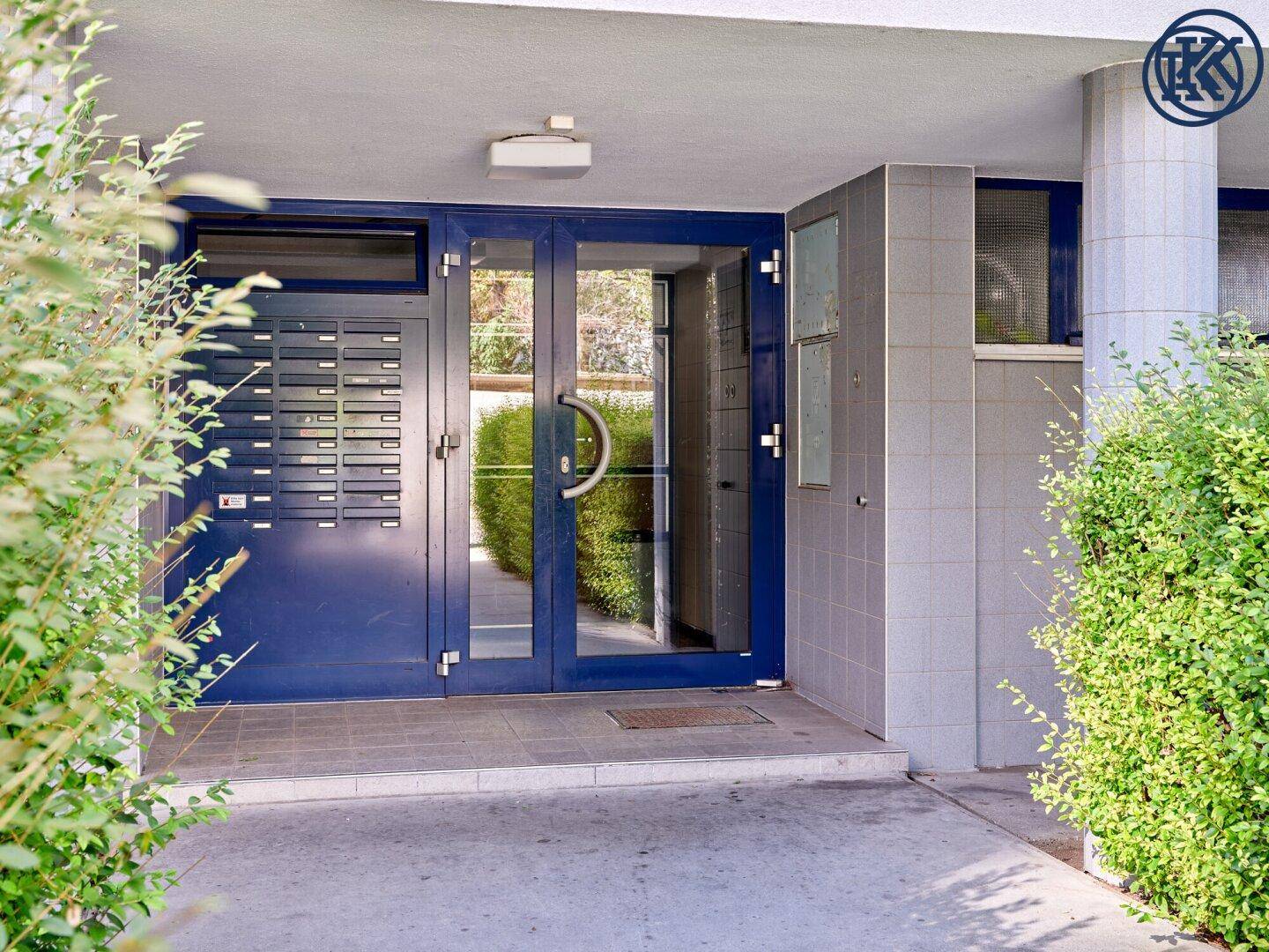 Eingangsbereich des Gebäudes mit blauen Türen, Briefkästen und umgebenden Büschen.